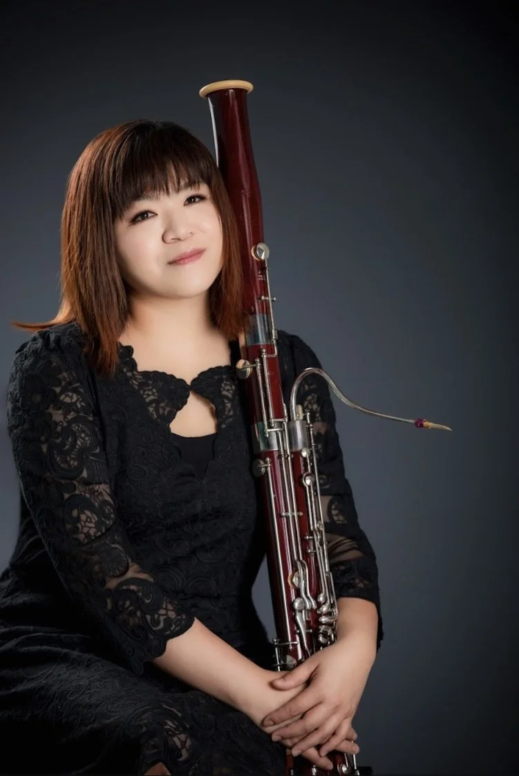 A woman with shoulder-length brown hair, wearing a black lace dress, is holding a bassoon, which is a woodwind instrument, against a dark gray background.