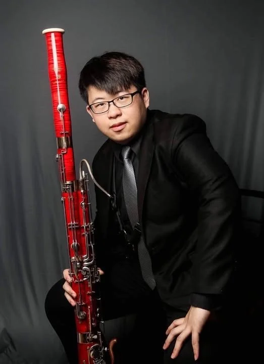 A young man in a black suit and glasses holding a large red bassoon, posing against a gray background.