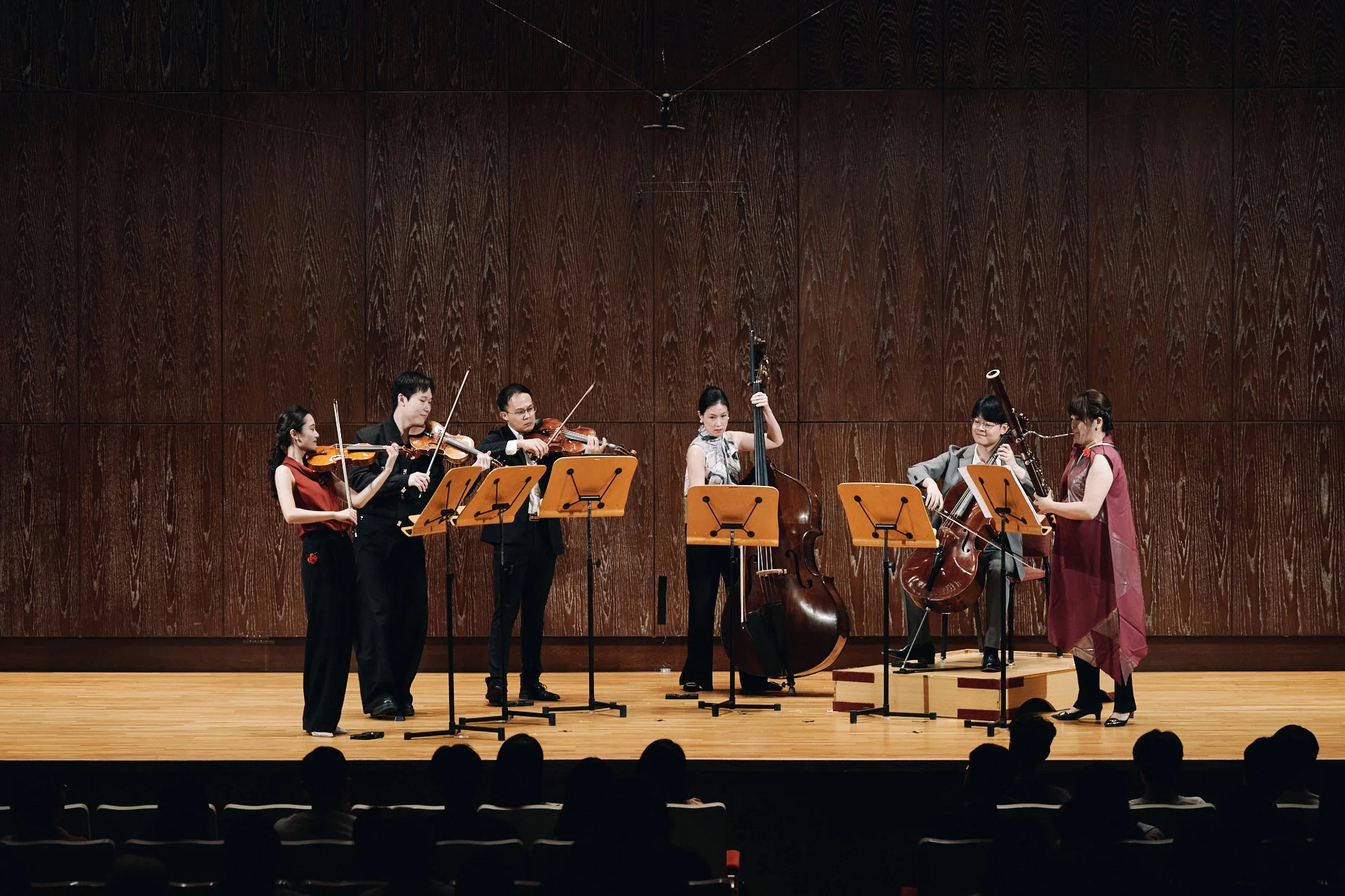 Group of six musicians performing on stage with three women and three men, playing violins and a double bass, in a concert hall with a wooden wall background.