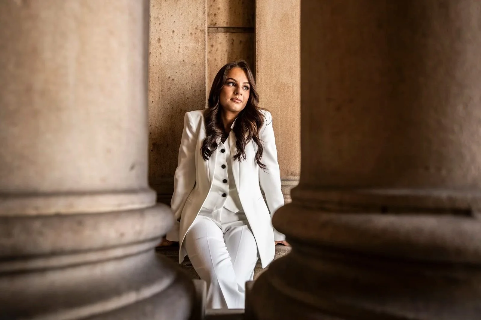 Woman in a white suit sitting on the floor between large stone columns in a marble building.