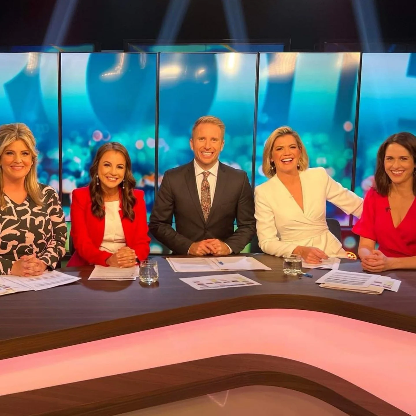 Five news anchors seated at a news desk during a broadcast, smiling and looking at the camera with papers and glasses of water in front of them.