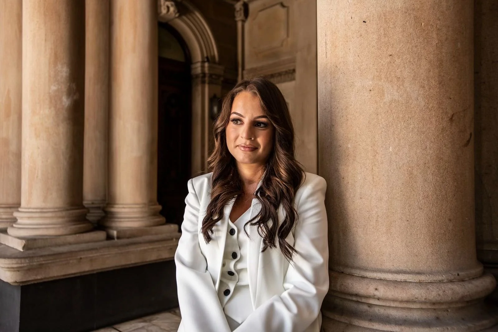 A woman with wavy brown hair, wearing a white blazer, standing next to large stone columns in a classical building.