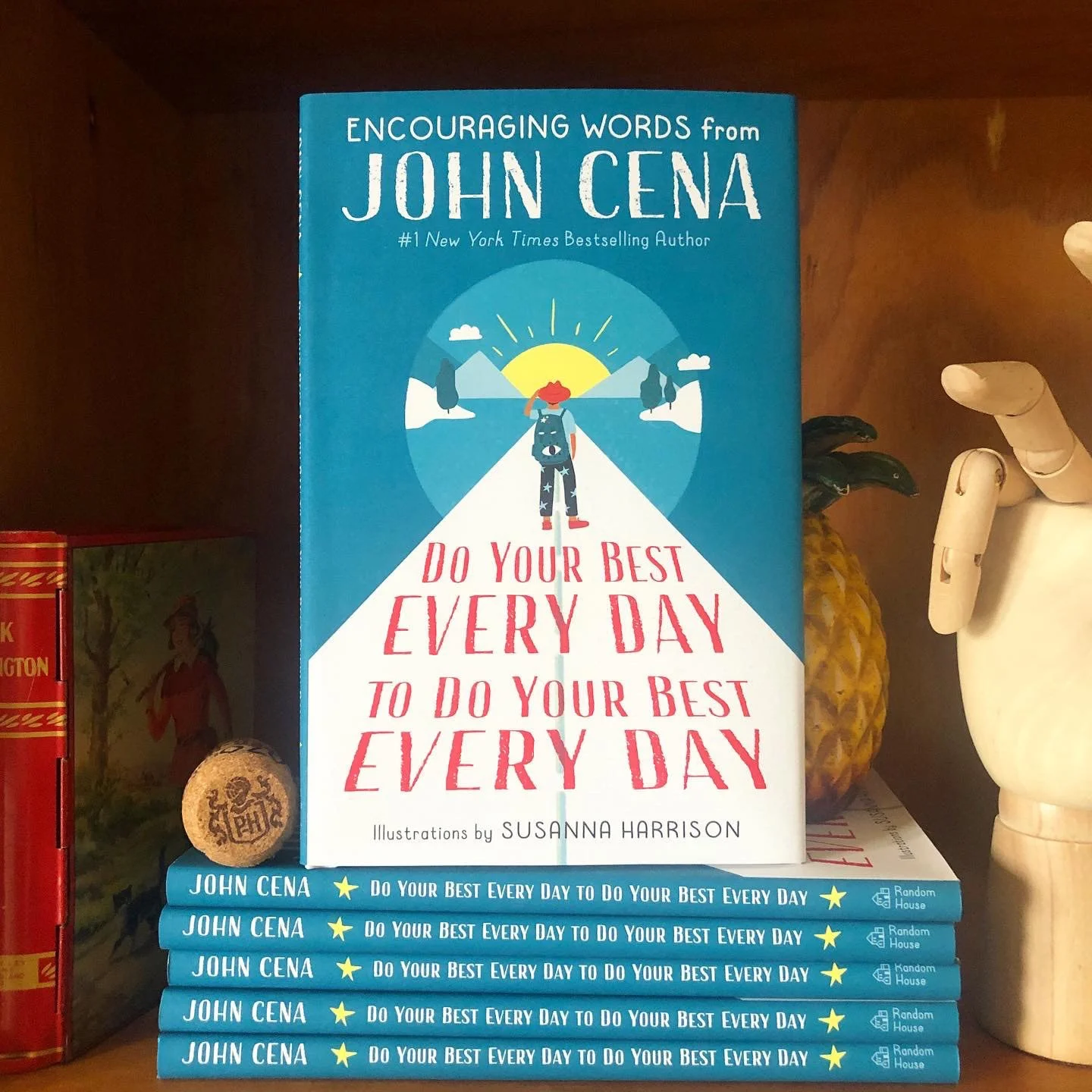 A stack of the book 'Do Your Best Every Day to Do Your Best Every Day' by John Cena, featuring illustrations by Paper Caravan founder Susanna Harrison. The blue books are displayed on a wooden shelf alongside an artist's wooden hand model