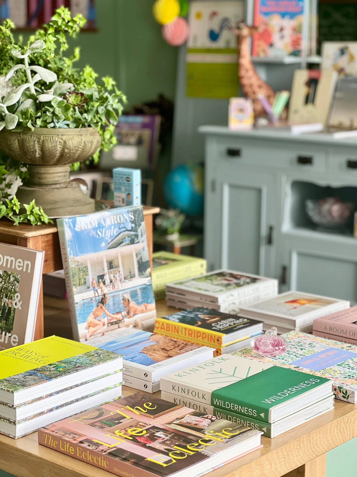 Interior view of Paper Caravan stationery shop in Newcastle, featuring a wooden table display of curated art books and colorful notebooks, with lush green plants and a vintage-style blue cabinet in the background.