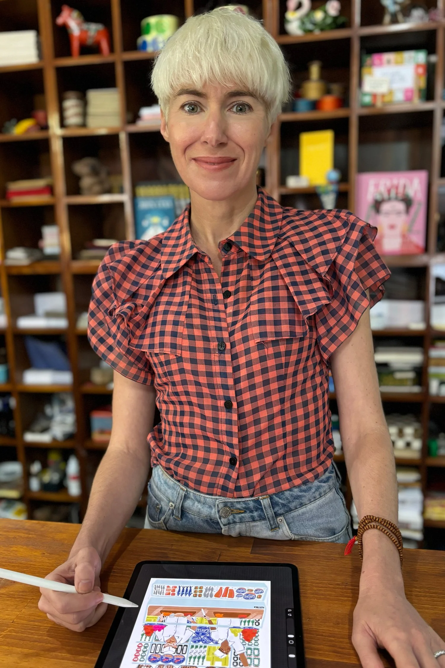 Susanna Harrison, professional illustrator and owner of Paper Caravan, standing in her Newcastle West shop with an iPad and Apple Pencil, surrounded by curated bookshelves.