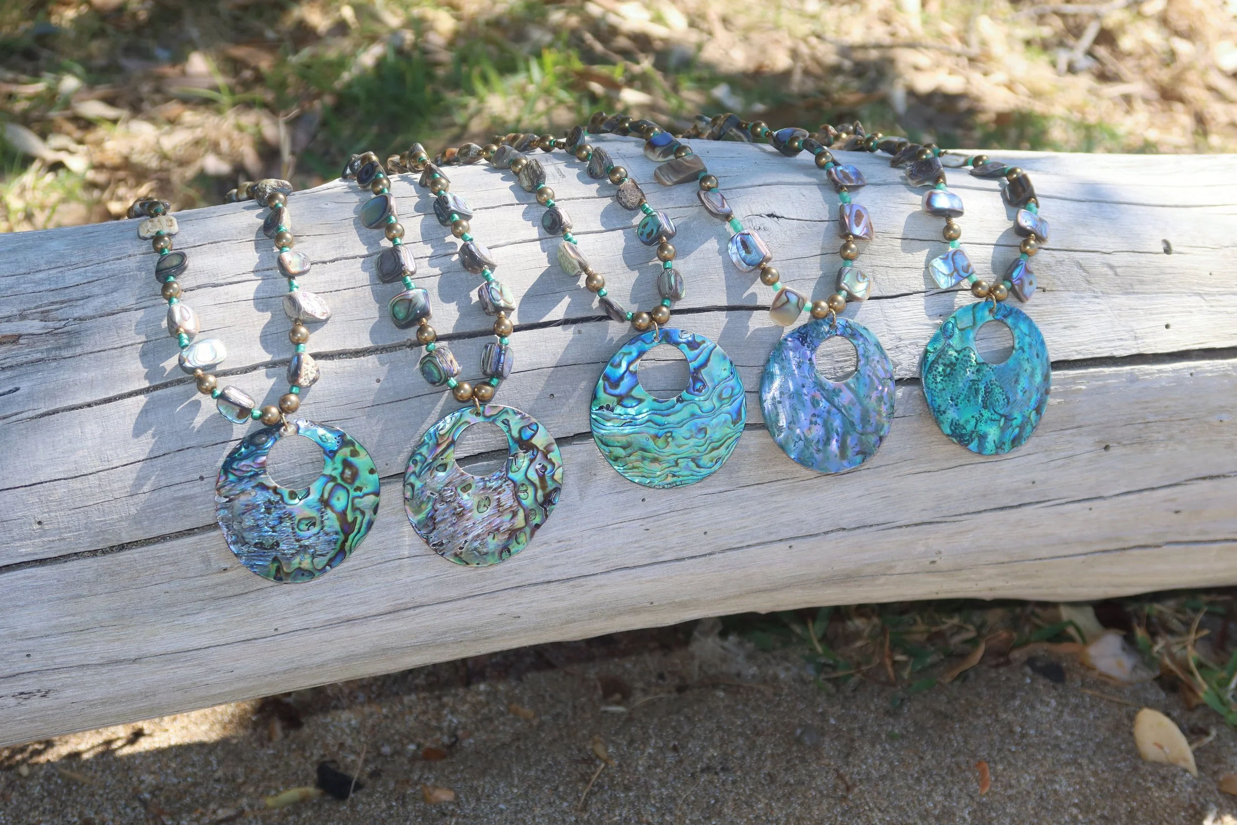Four necklaces with large, round, iridescent shell pendants and smaller metallic beads are laid out on a weathered wooden log outdoors, with grass and soil in the background.