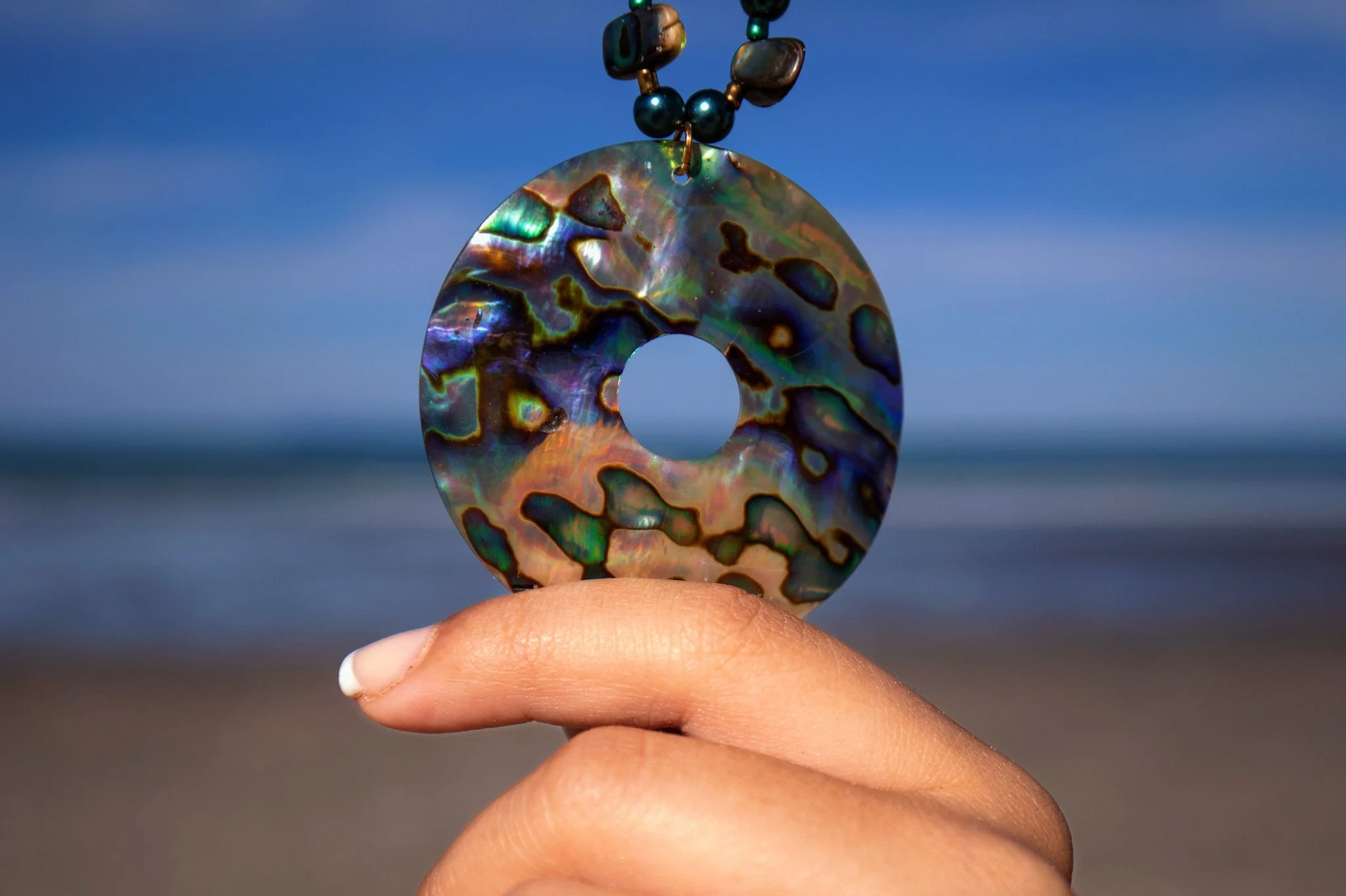 A person is holding an iridescent, multicolored circular object with a hole in the center, resembling a shell or a gemstone, against a clear blue sky and ocean background.