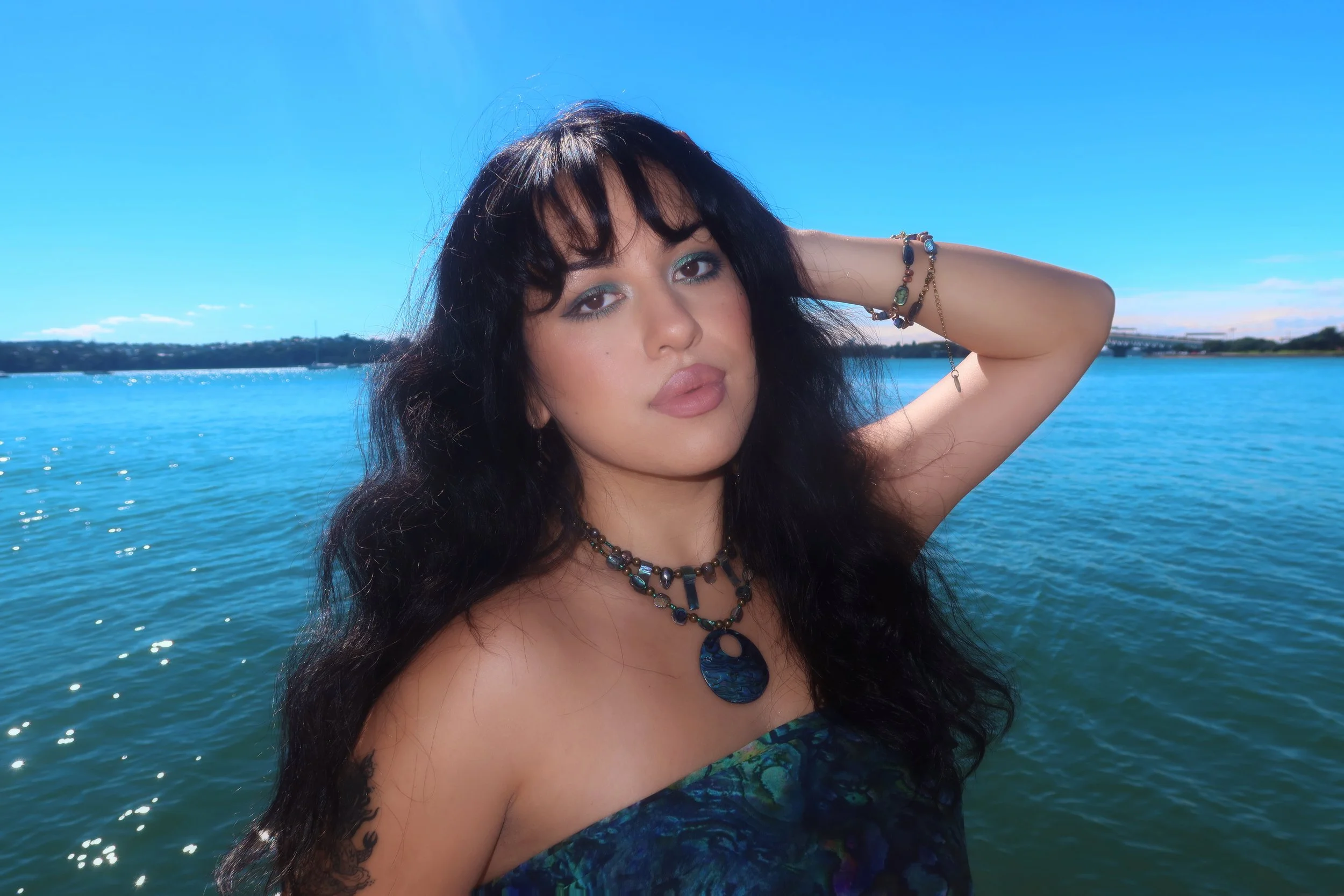 A woman with long black hair and jewelry posing outdoors near a body of water under a bright blue sky.