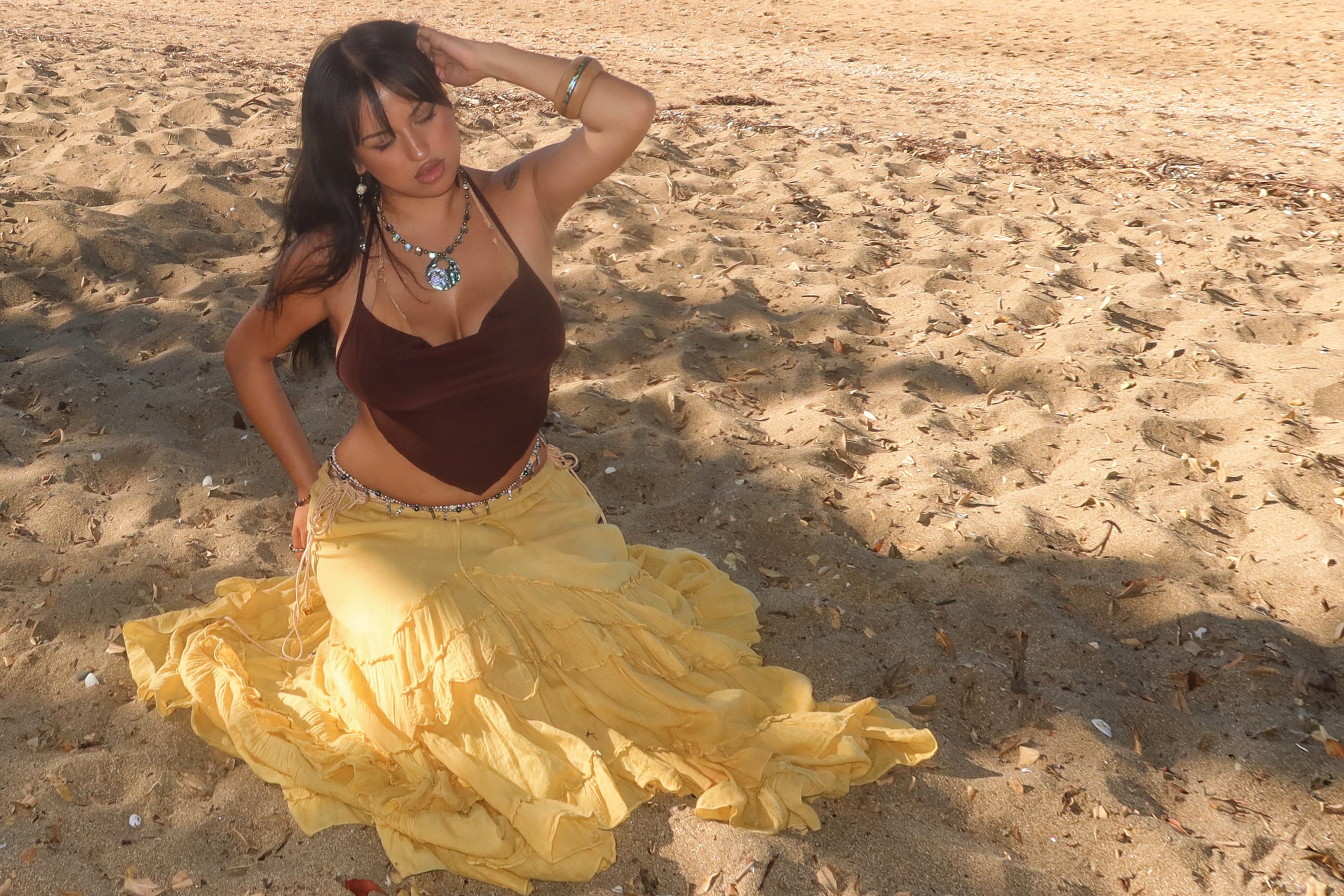 A woman with dark hair, wearing jewelry, a dark sleeveless top, and yellow skirt, poses on a sandy beach with seashells and footprints, with her eyes closed and one arm raised to her head.