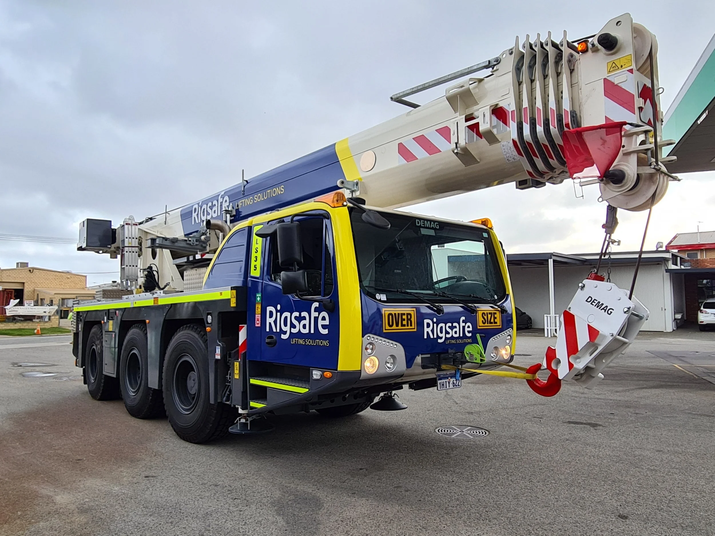 A mobile crane truck with yellow and blue coloring, labeled 'Rigsafe' and 'Lifting Solutions'. The crane's boom extends upward, and a red and white striped counterweight is attached to the end. High visibility DG3 striping and Asset Number
