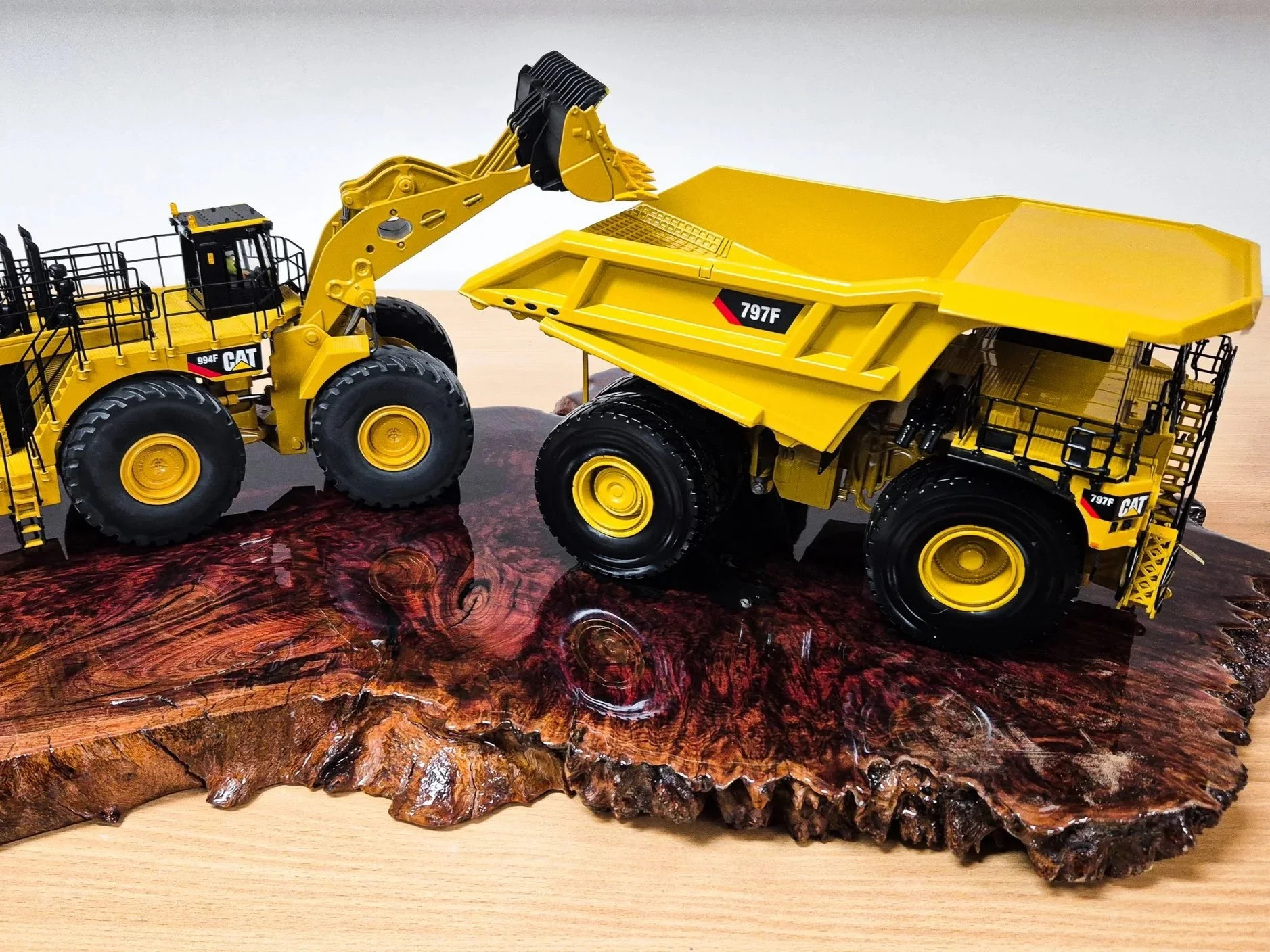 A detailed scale model of a yellow Caterpillar front loader placed on a wooden surface with a large piece of polished wood beneath it. Custom mining gifts