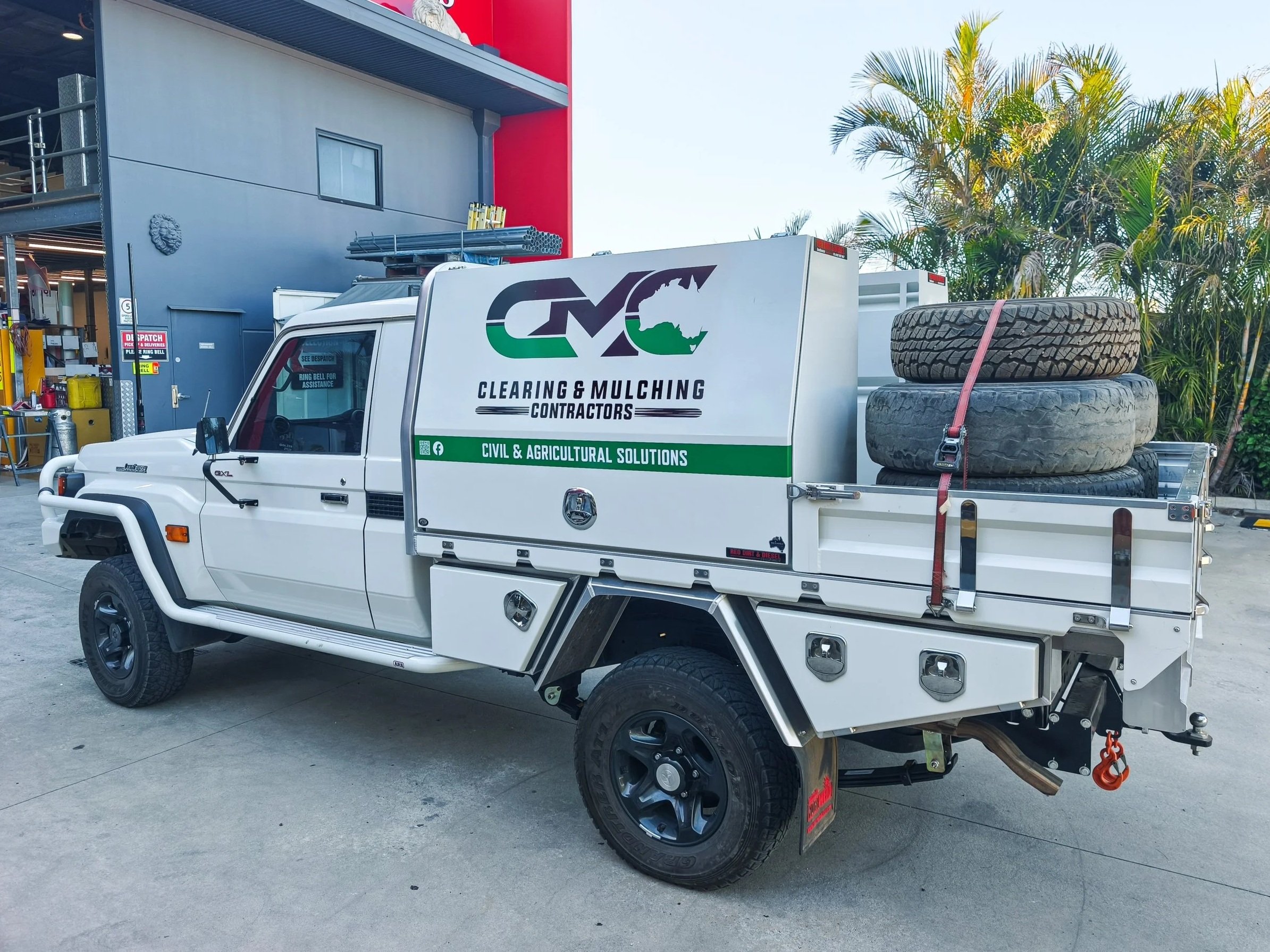 A white service truck with a logo and text for a clearing and mulching contractors business, parked outside a building with a red and gray facade and palm trees in the background. The truck has large tires, a flatbed with tires, and equipment on the side.