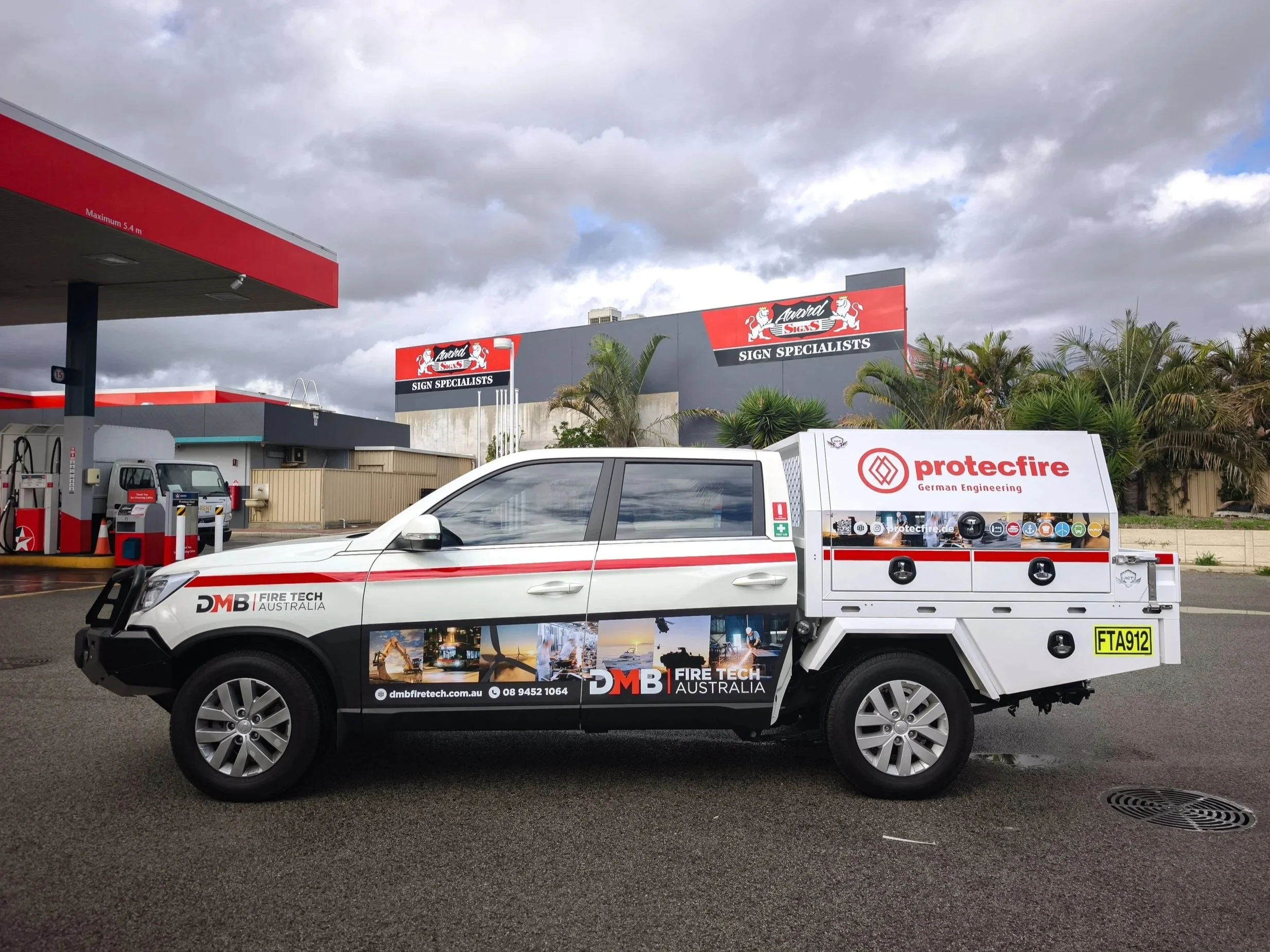 A white utility vehicle with branding for DMB Fire Tech Australia parked at a gas station. The vehicle advertises fire technology services with website, contact number, and promotional images. In the background, there are commercial buildings with signs for 'Award Signs' and 'Protec Fire,' palm trees, and a cloudy sky.