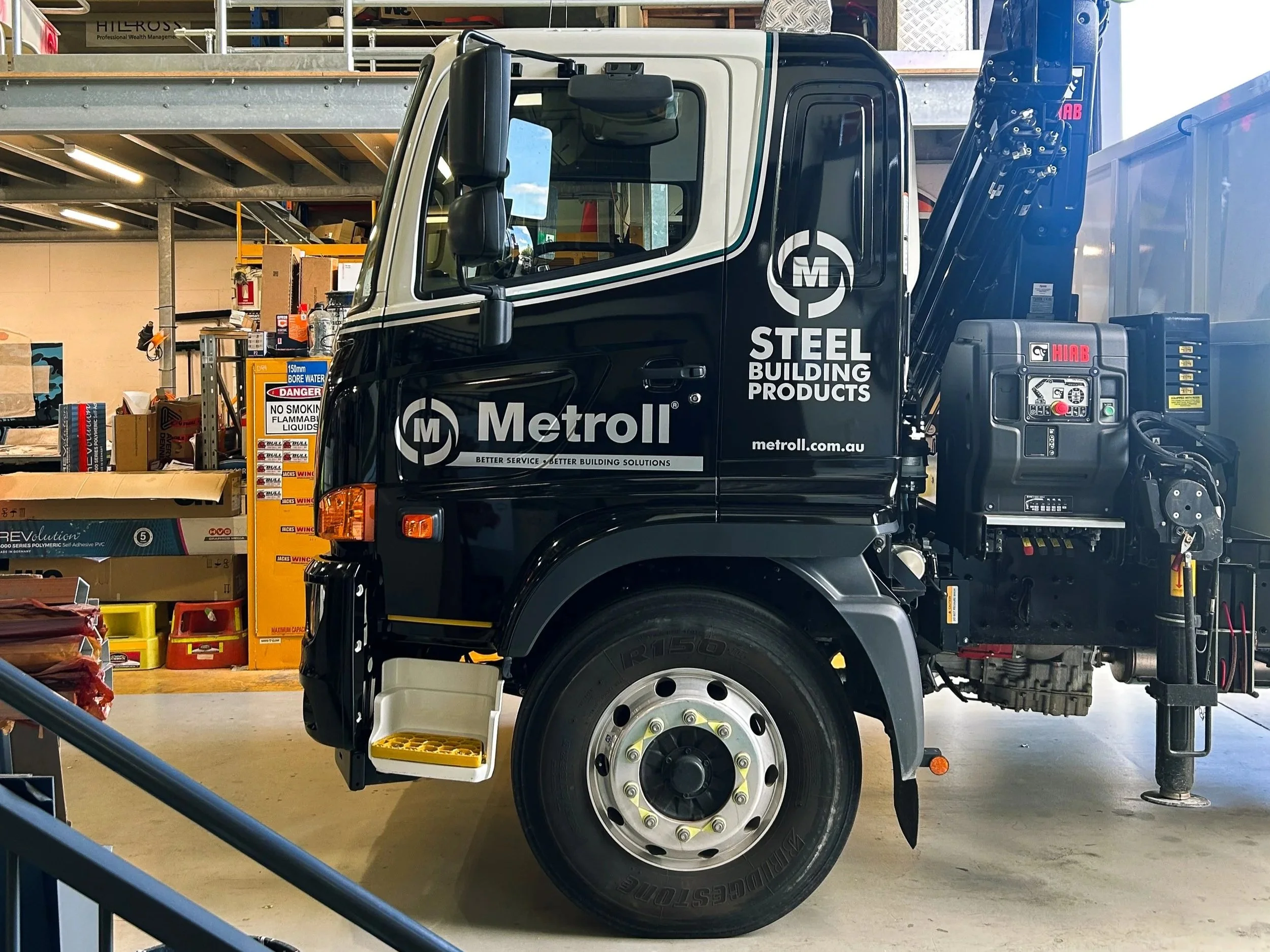 A black and white truck inside a warehouse with shelves, boxes, and tools. The truck has the logo "Metroll" and the words "Steel Building Products" on the door. The truck is equipped with a hydraulic arm and control panel.