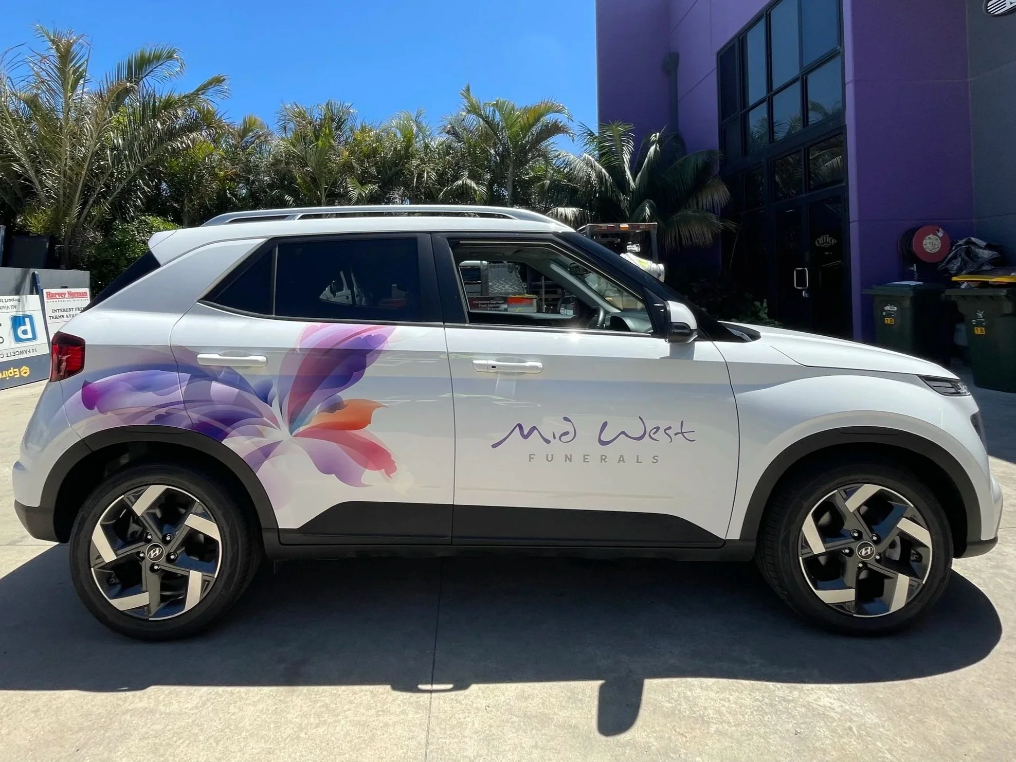 A white vehicle with floral graphics and the text "Mid West Funerals" parked outside a building with purple walls, green trees, and trash bins nearby.