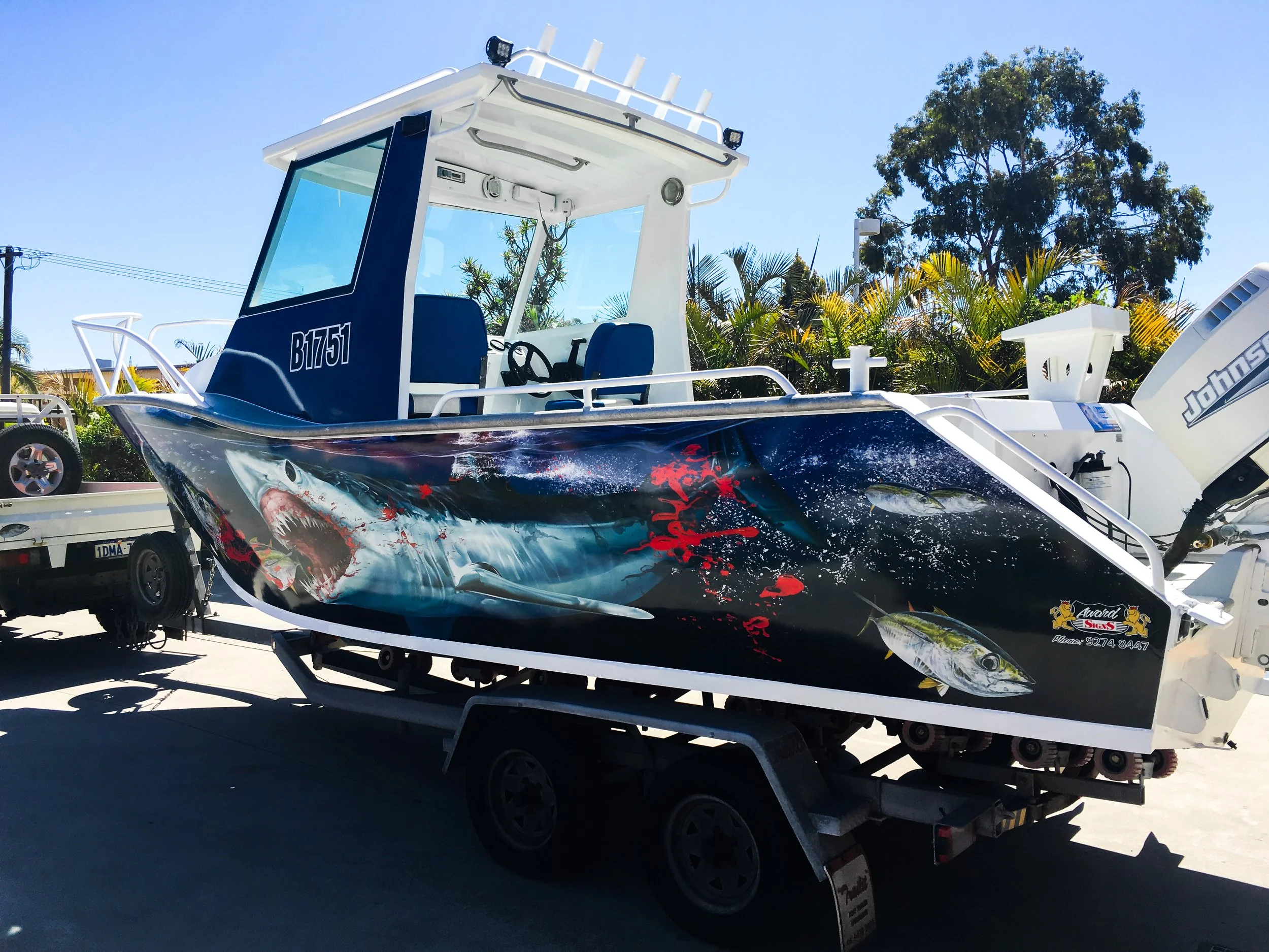 A boat with a shark graphic and aggressive design on its side is on a trailer in an outdoor lot. The boat has a blue and black color scheme with an elaborate graphic of a shark with red accents. The boat's interior has blue seats and a steering wheel, and the outboard motor at the back displays the brand "Johns". There are trees and a utility pole in the background under a clear blue sky.