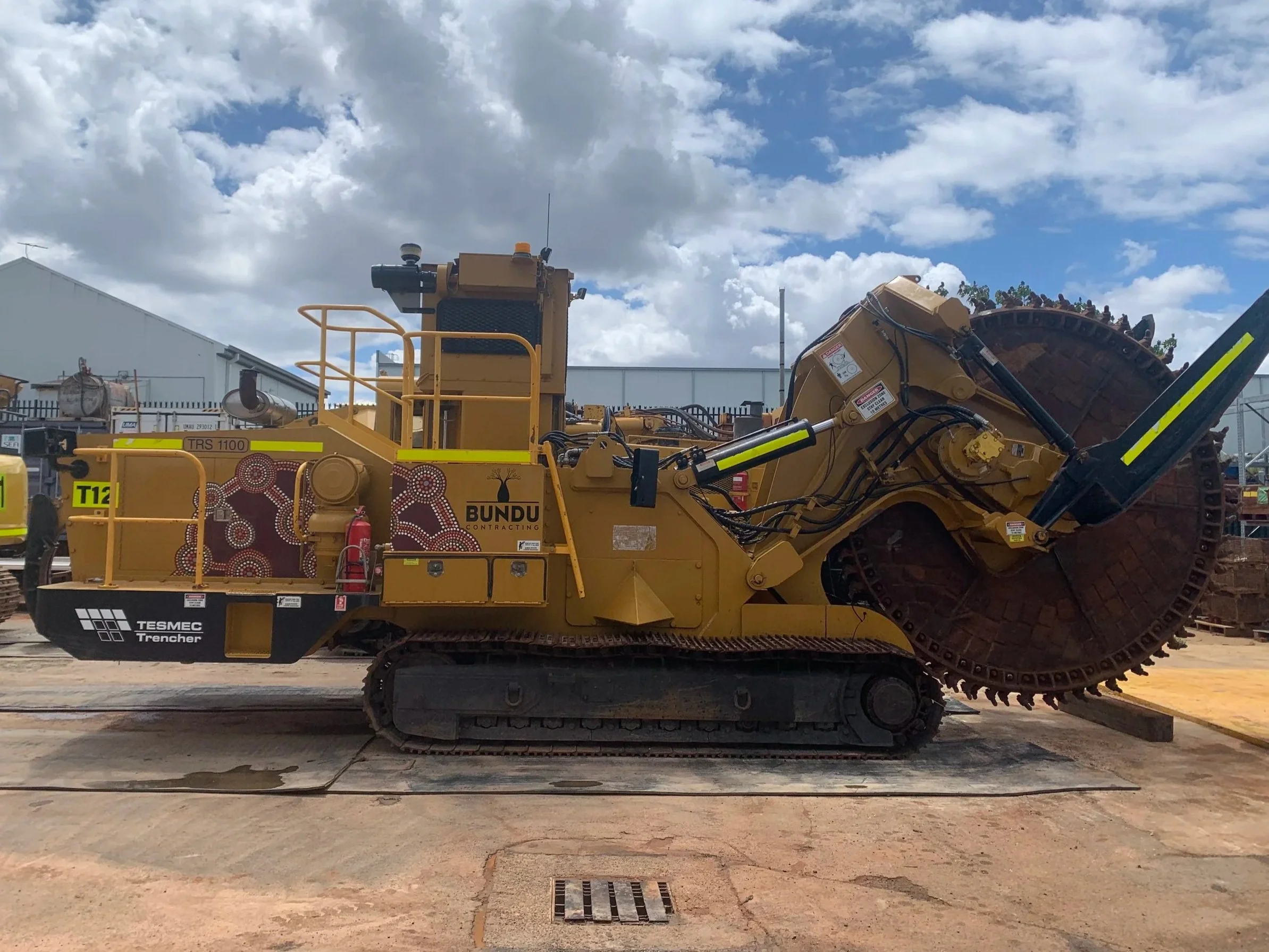 A large yellow trencher mining machine, with a tracked base and a cutting chain at the front, branded with 'BUNDU' and 'TESMEC'.  Industrial Machinery Signage and Branding with High Visibility DG3 Striping