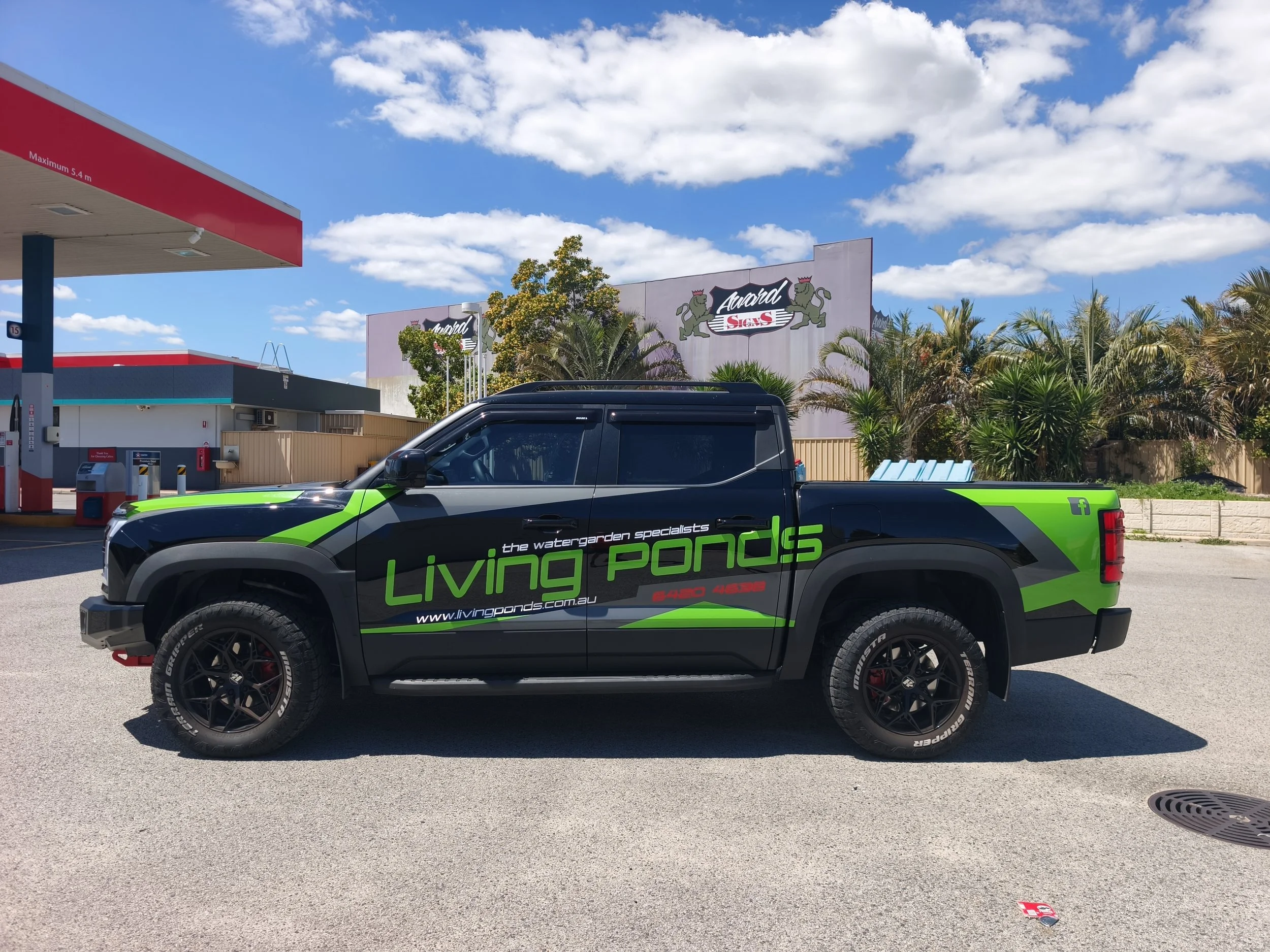 Black and green pickup truck parked at a fuel station with a sign for Living Ponds and a blue sky with white clouds in the background.