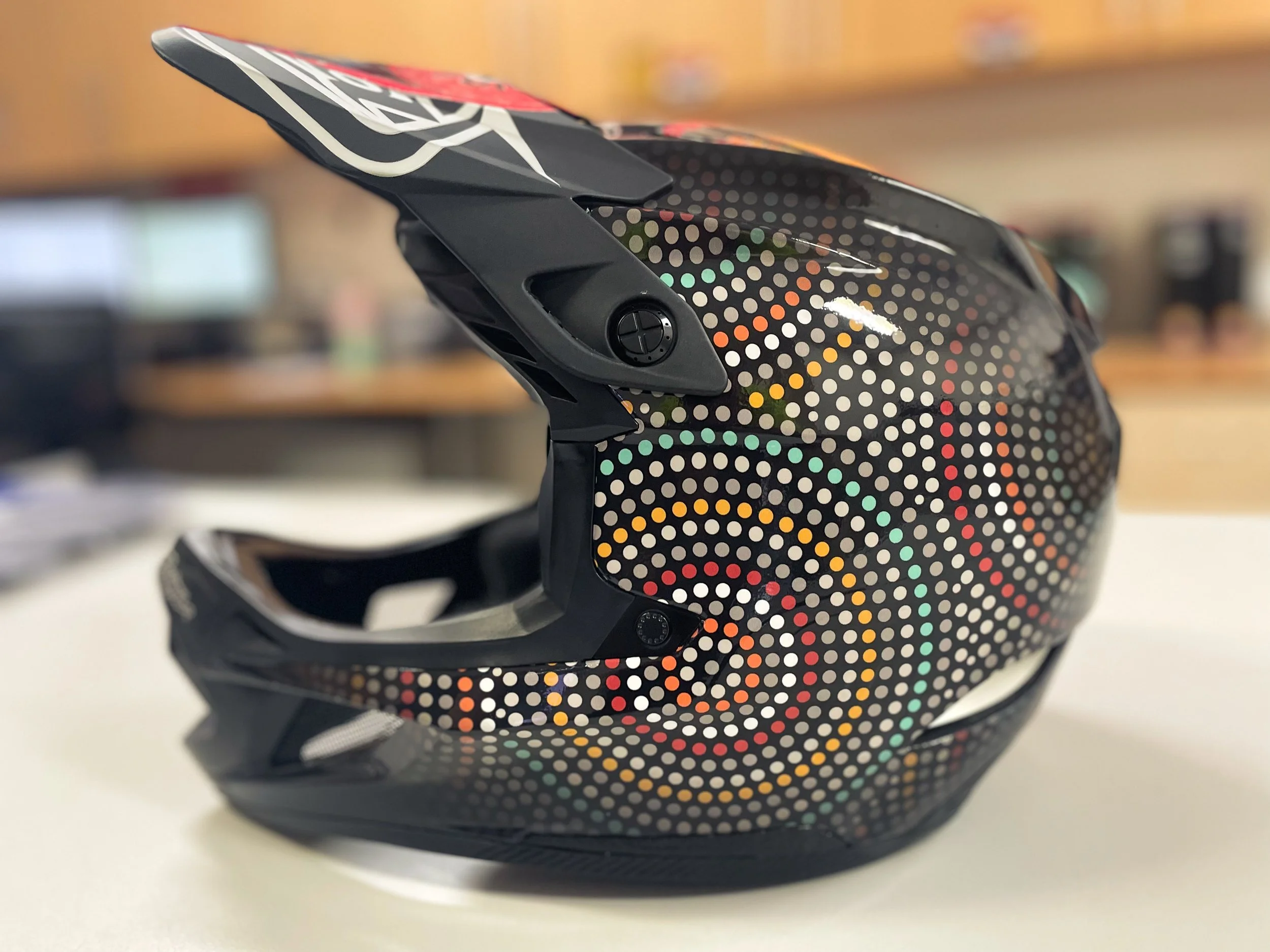 A black motorcycle helmet with a colorful dotted swirl pattern and a black visor, resting on a white surface.