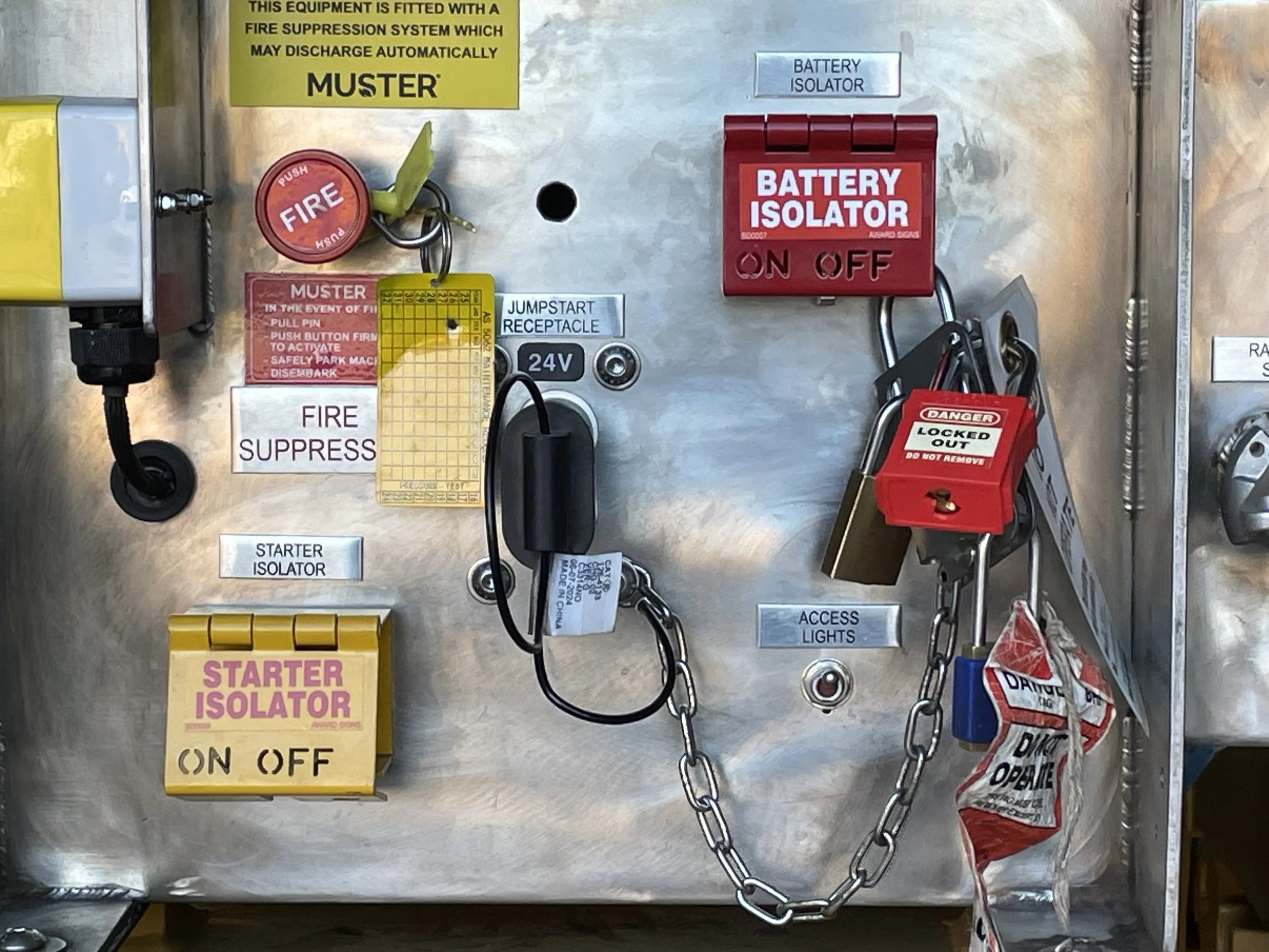 Panel with various safety and equipment signs and devices, including a red fire alarm button, a yellow label, a red battery isolator switch, and a black lockout device, mounted on a metallic surface.