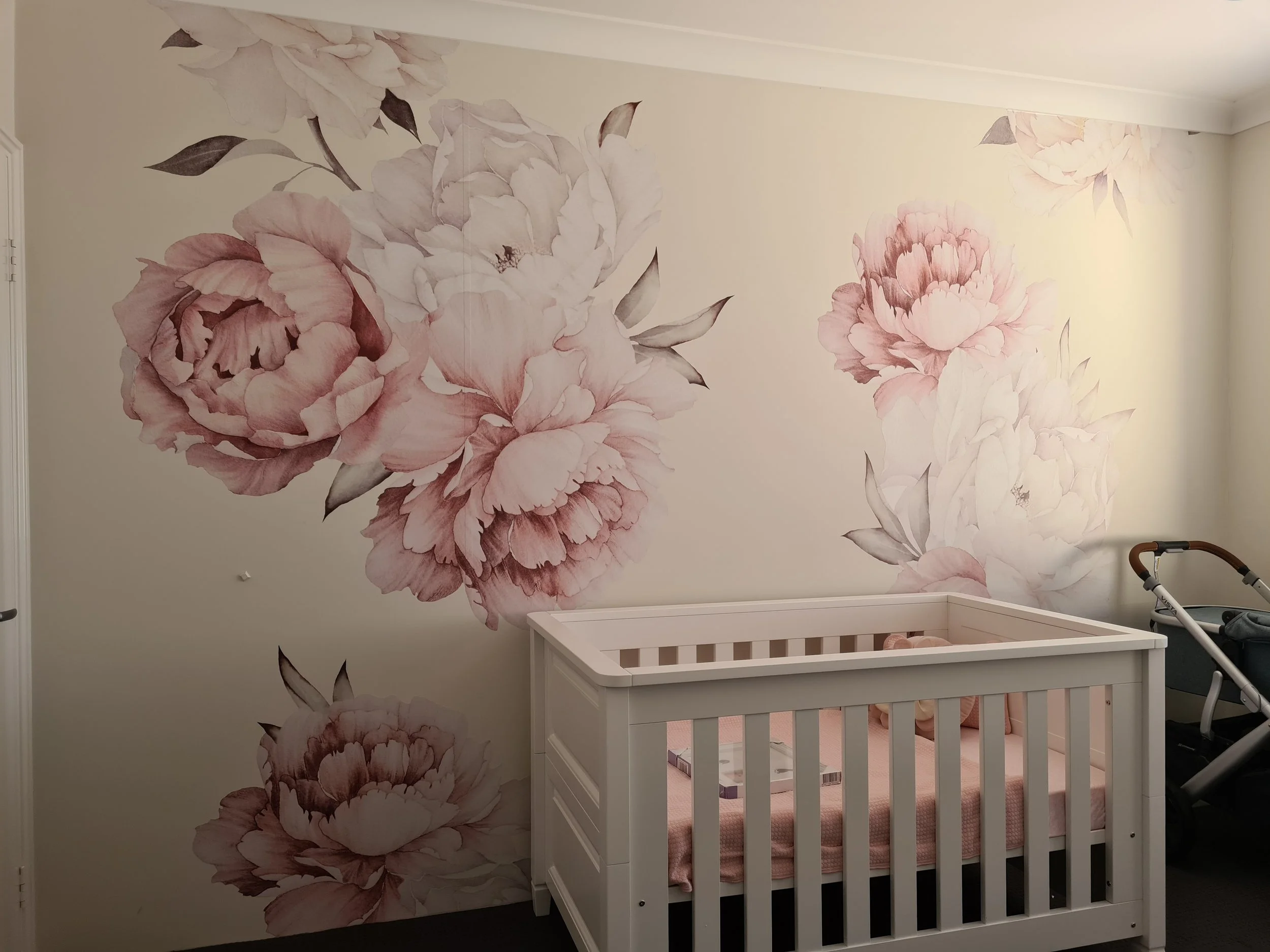 A nursery with a floral wall mural featuring large pink and white peonies. There is a white crib with a pink blanket inside and some baby items below it. A stroller is partially visible on the right side.