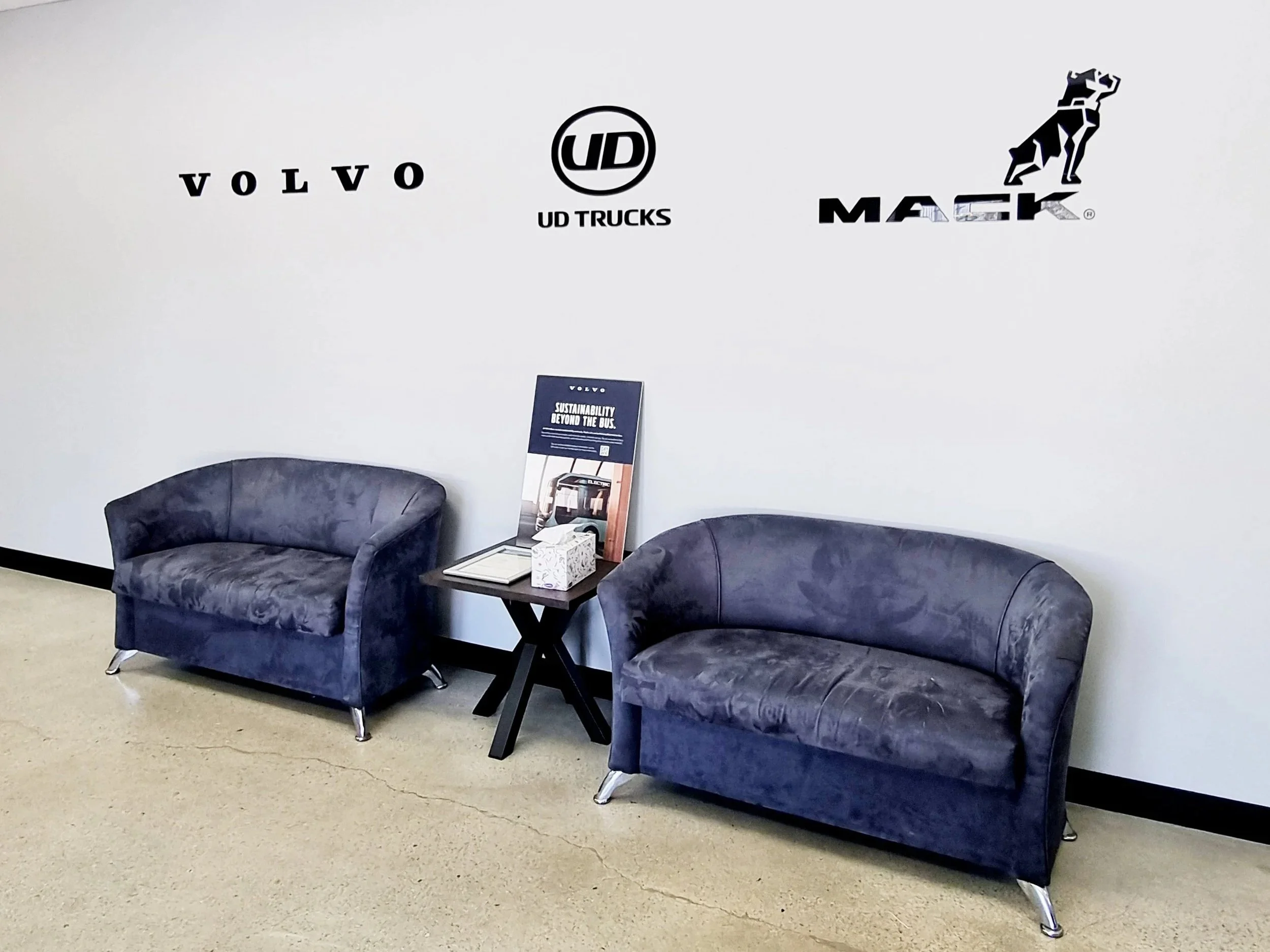 Waiting area with two black couches, a small table holding brochures, and acrylic letter branding on the wall including Volvo, UD Trucks, and Mack logos. Truck Centre WA. Reception signage