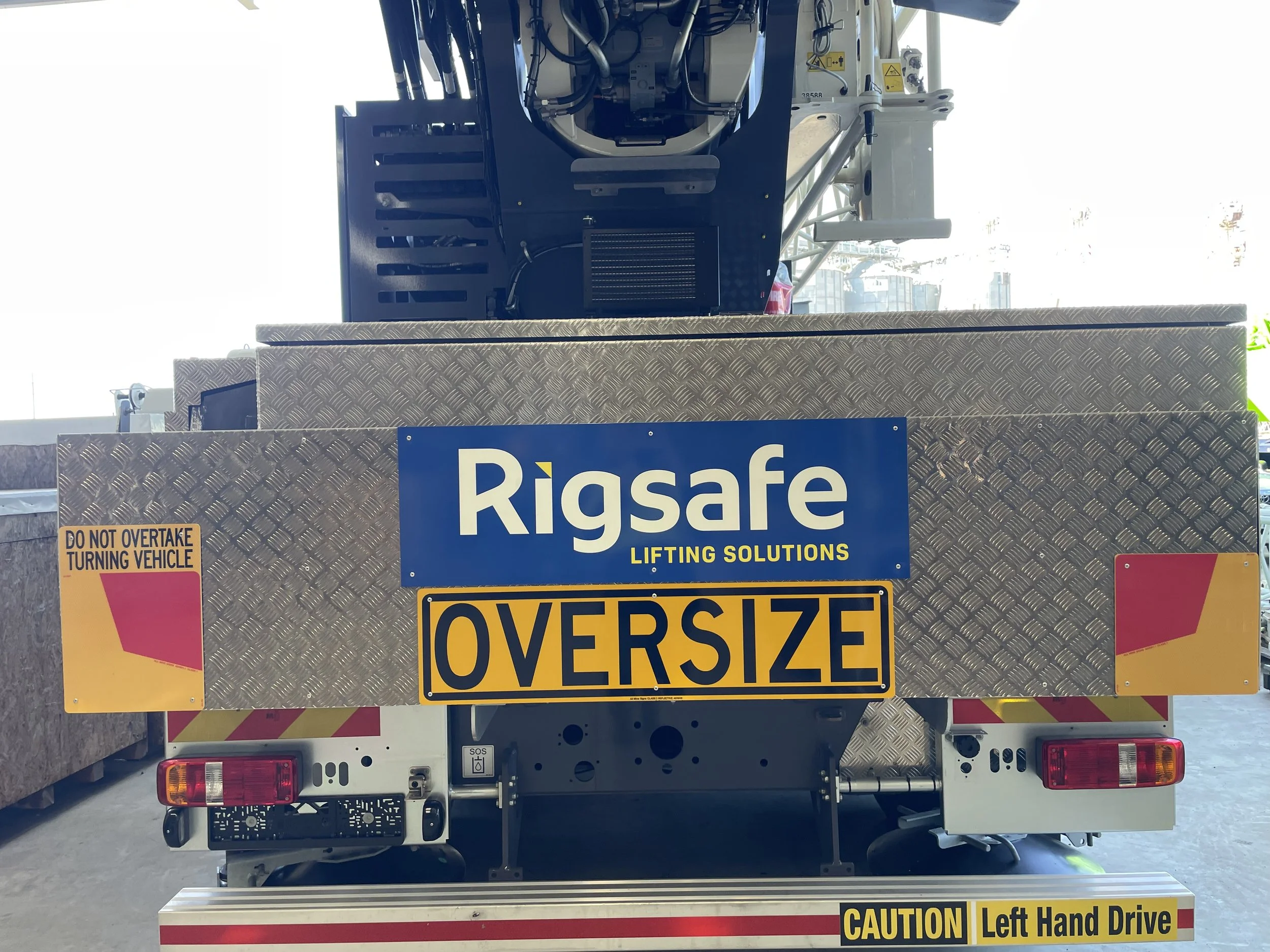 Back of a utility vehicle with a sign that reads 'RigSafe Lifting Solutions' and 'Oversize,' with warning labels and safety instructions, and equipment mounted on the top.