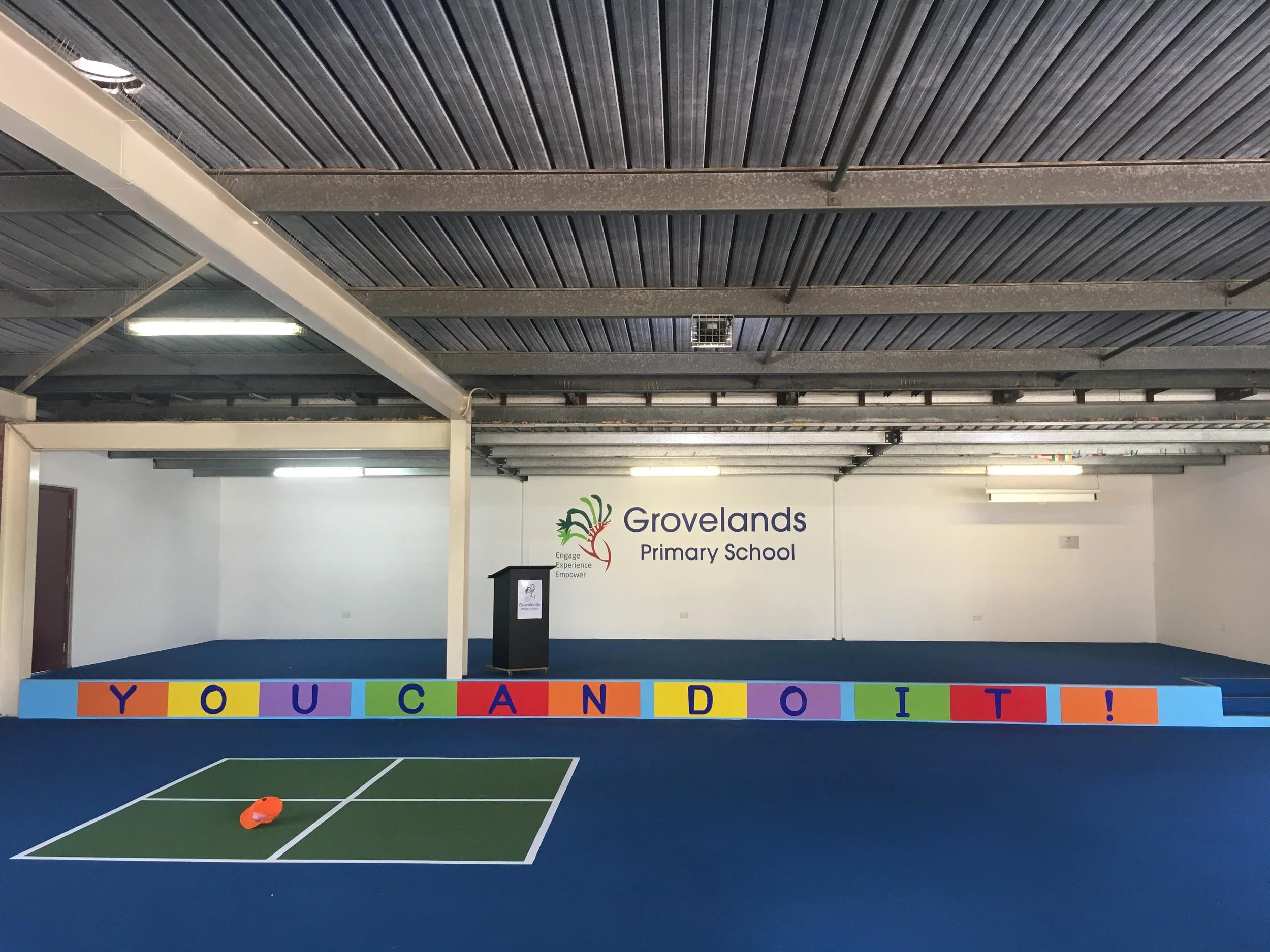 Indoor sports court with a colorful banner saying 'You Can Do It!' and a sign for Grovelands Primary School on the wall. Education Signage
