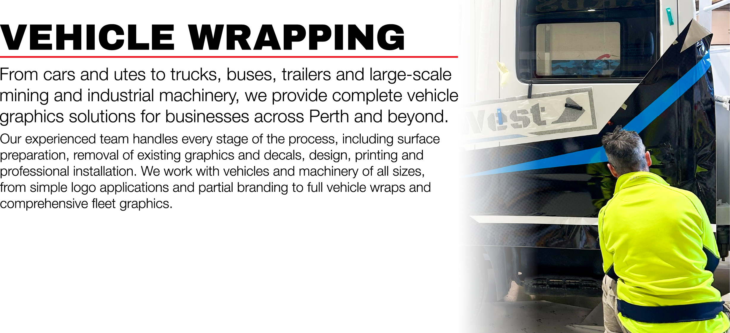 A person in a bright yellow safety jacket is applying vehicle graphics to a large black vehicle, using tools and with graphics already partially installed.