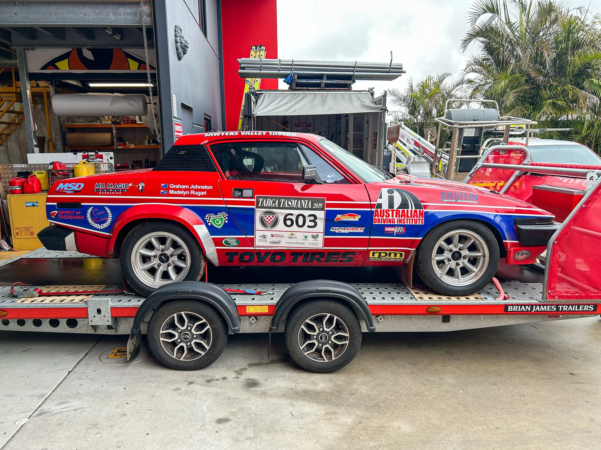 A red race car with various sponsor logos and driver names, loaded on a trailer with a metal platform and planks, surrounded by equipment and palm trees in the background.