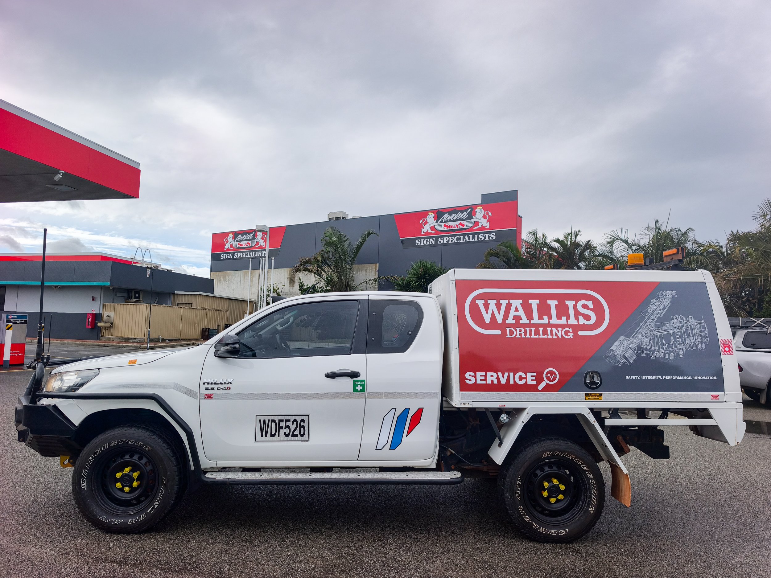 A white pickup truck with graphics and the license plate WDF526 parked in a lot. The truck has a service body for Wallis Drilling with tools and safety equipment. In the background, there are buildings with signage for Award Signs and Wallis Sign Specialists, as well as trees and cloudy skies.