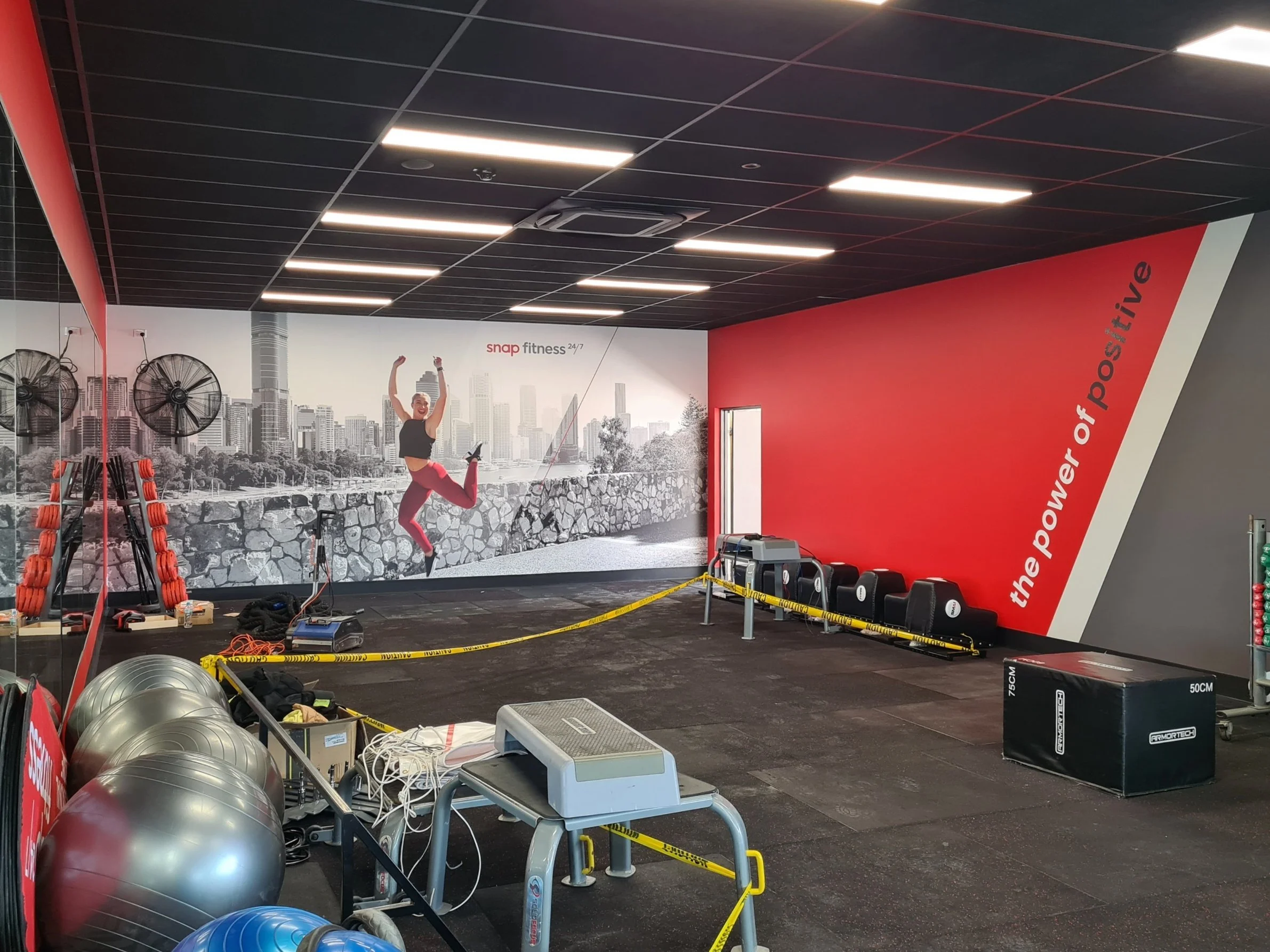 A gym workout area with a large mural of a city skyline and a woman in fitness attire jumping with arms raised. Gym equipment includes exercise balls, weights, and weight racks. The ceiling has black panels with integrated lighting, and the area is bordered with yellow caution tape.