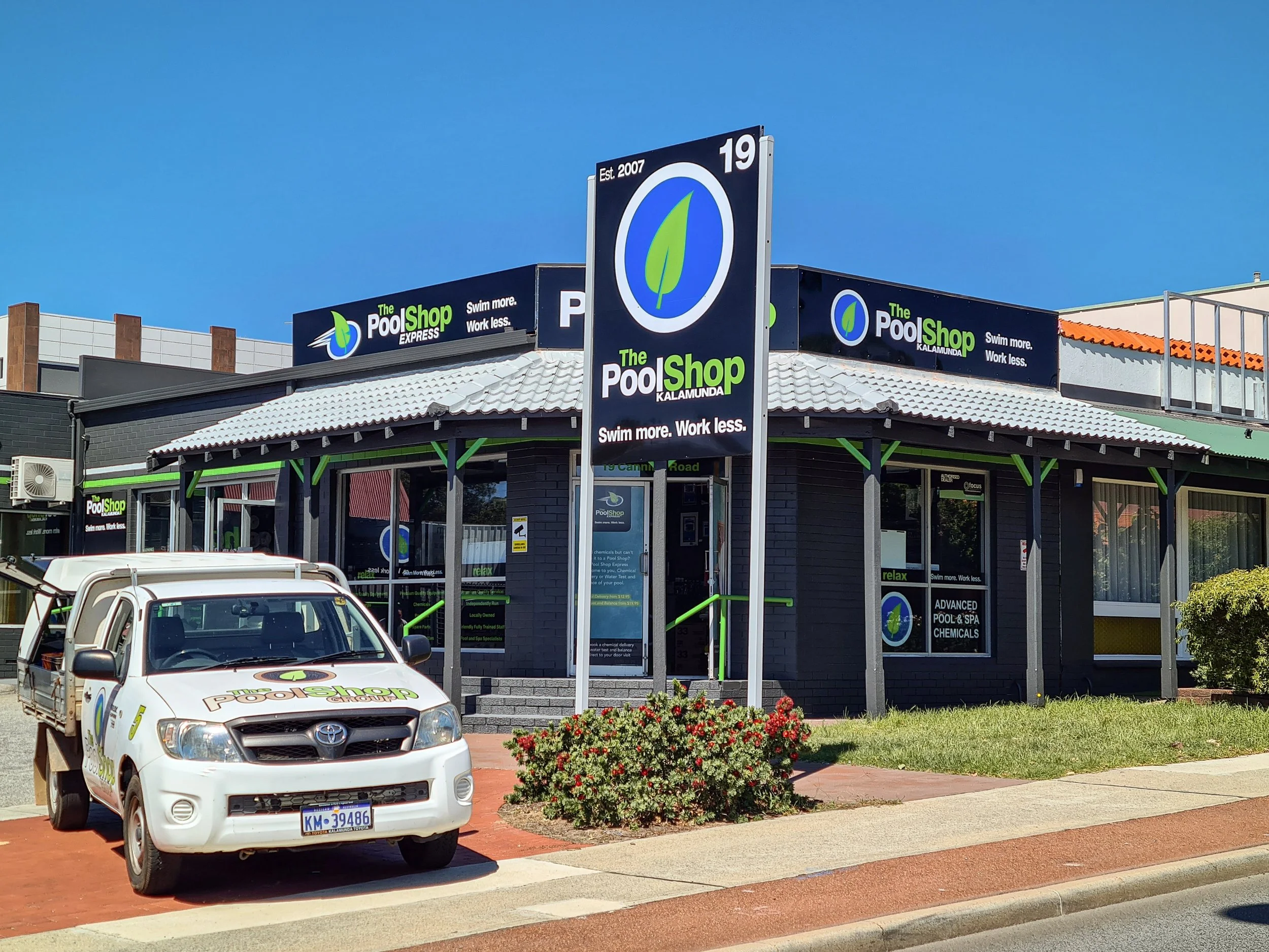 Exterior of a store named 'The Pool Shop Kalamunda' with signs that read 'Swim more. Work less.' and 'Advanced Pool & Spa Chemicals.' A white Toyota Hilux with the store's logo on the bonnet is parked in front. Business Building and Vehicle Signage