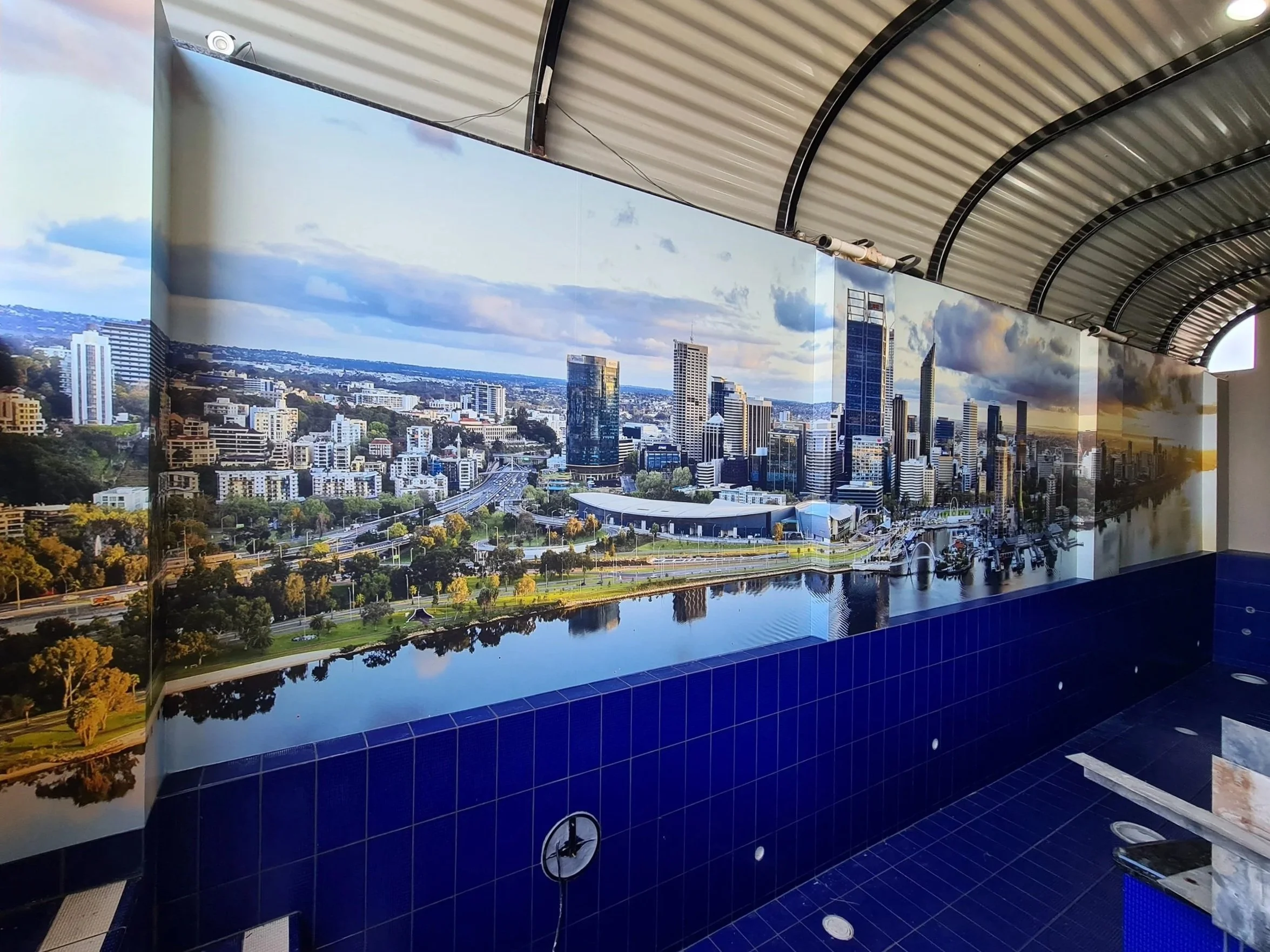 A large wall mural of a city skyline with tall buildings, roads, and a river, displayed on the interior wall of a pool area with a curved metal roof ceiling. Personalised wall signage
