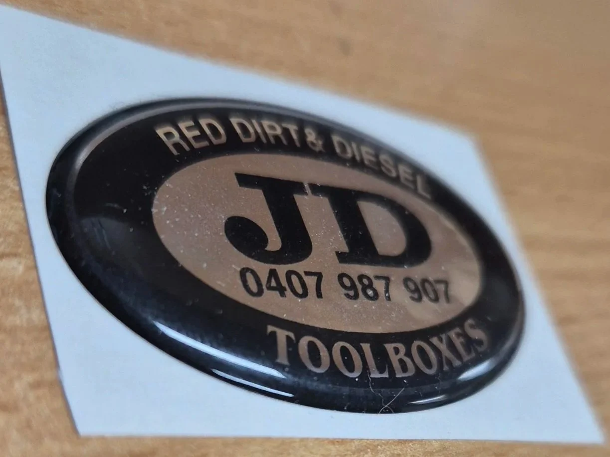 Domed sticker decal with a black and beige logo reading 'JD TOOLBOXES' and 'RED DIRT & DIESEL' along with a phone number. Custom decals