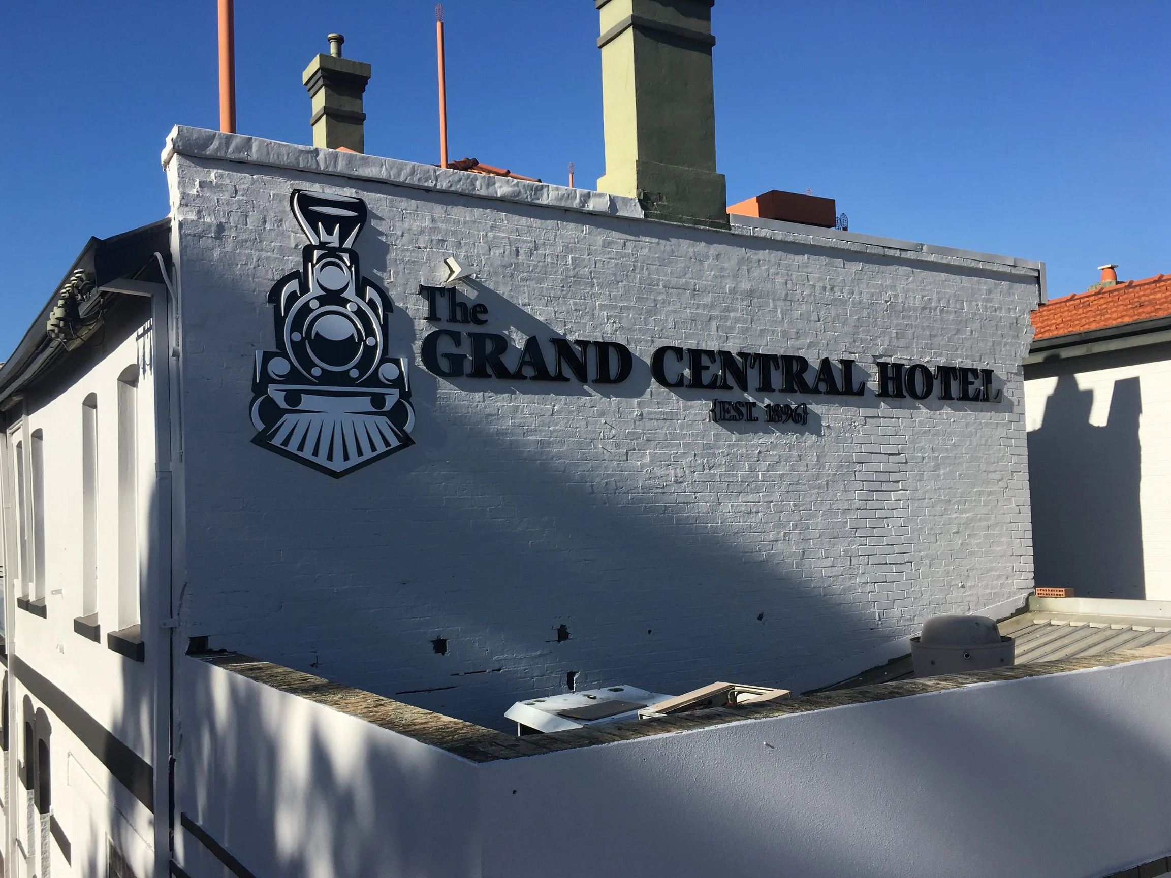 A white brick building with edged decals that reads 'The Grand Central Hotel' in Midland and features a stylized train logo. The building has two tall chimneys and a red tiled roof, set against a clear blue sky.