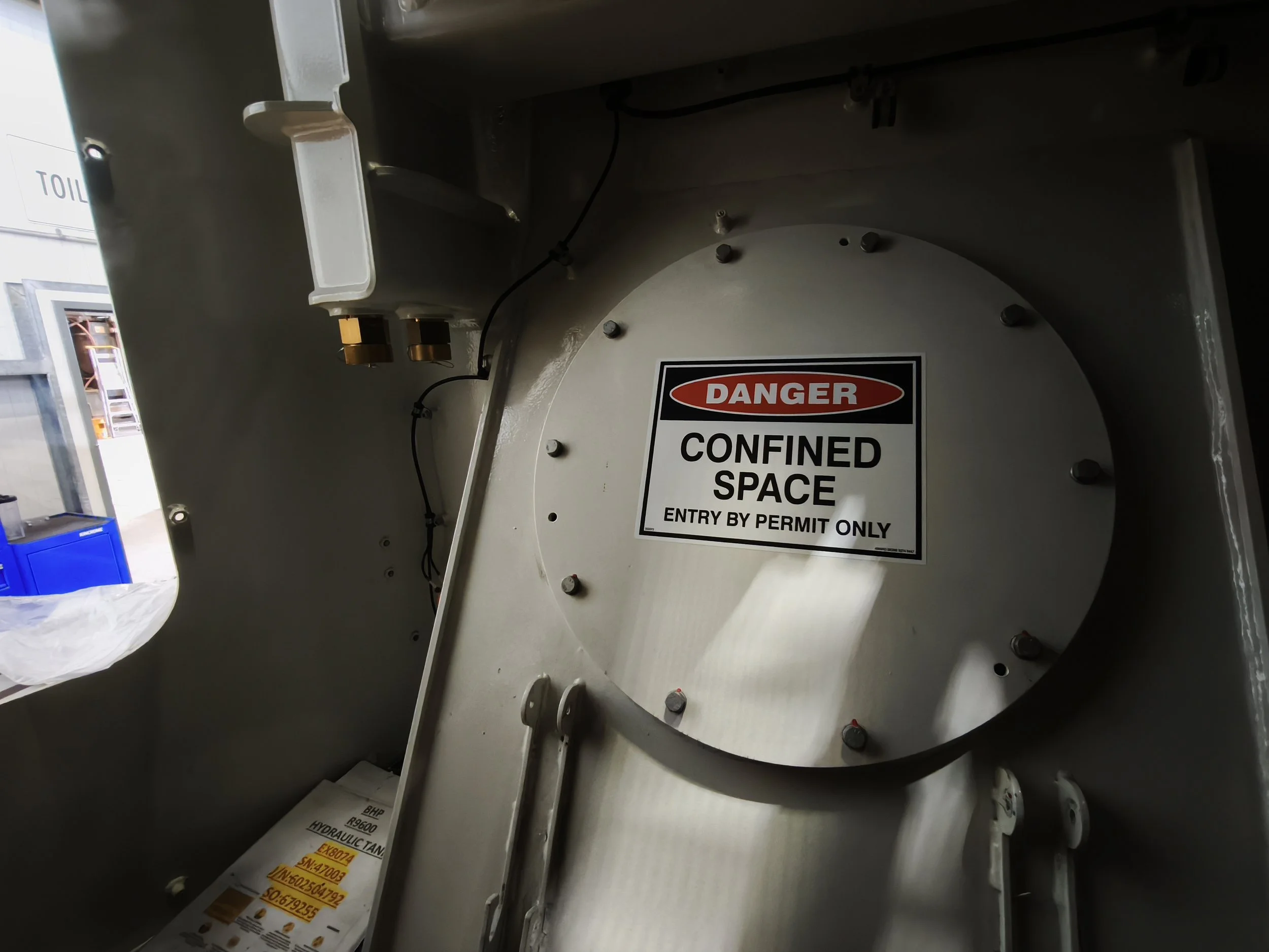 Close-up view of a metallic confined space with a warning label that reads 'Danger Confined Space Entry by Permit Only'. The space contains wires, bolts, and a white component mounted on the side.