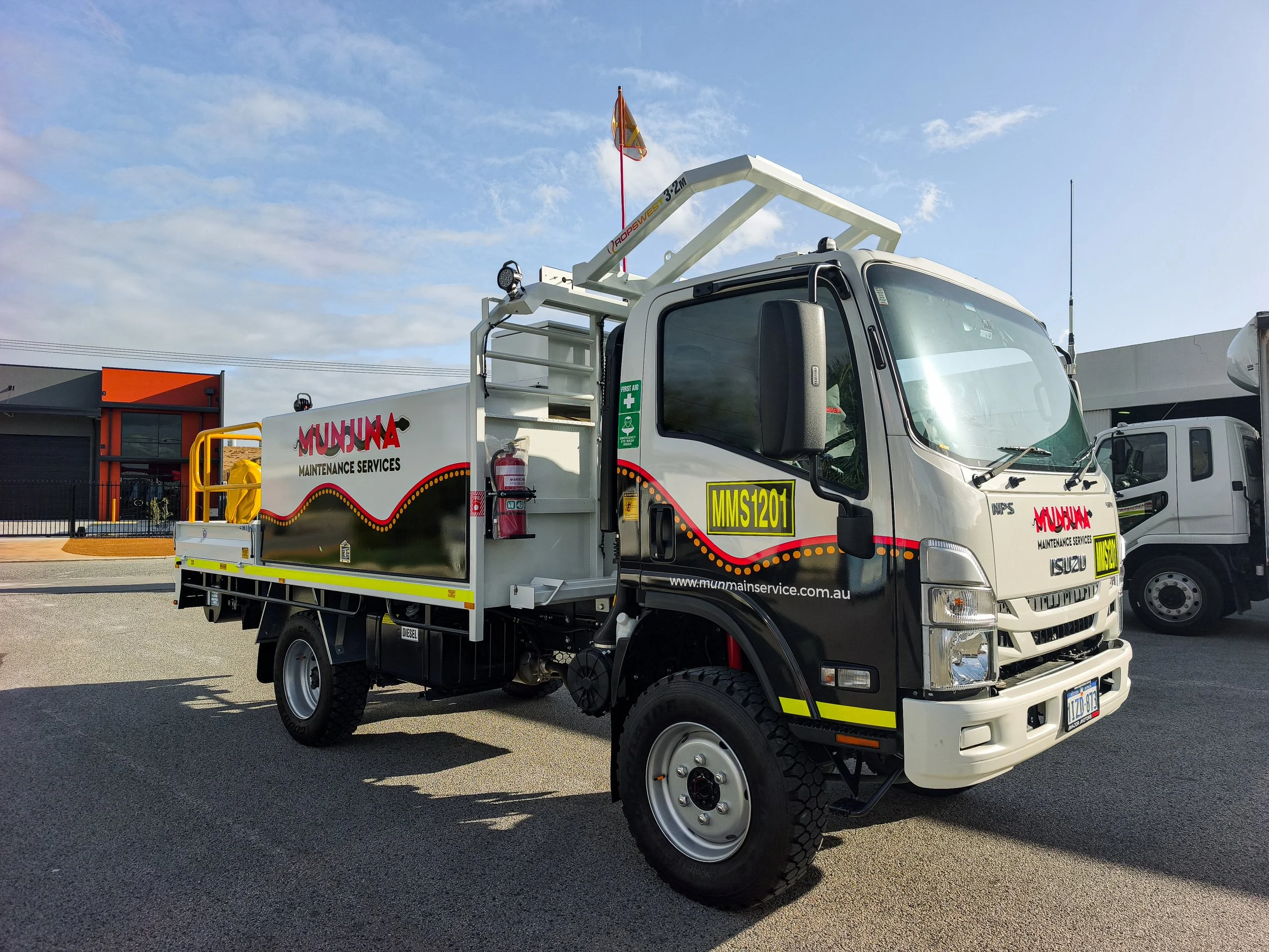 A service truck labeled 'Munmua Maintenance Services' parked outdoors under a partly cloudy sky. The truck has a cherry picker lift, first aid kit, fire extinguisher, and various equipment on its flatbed.