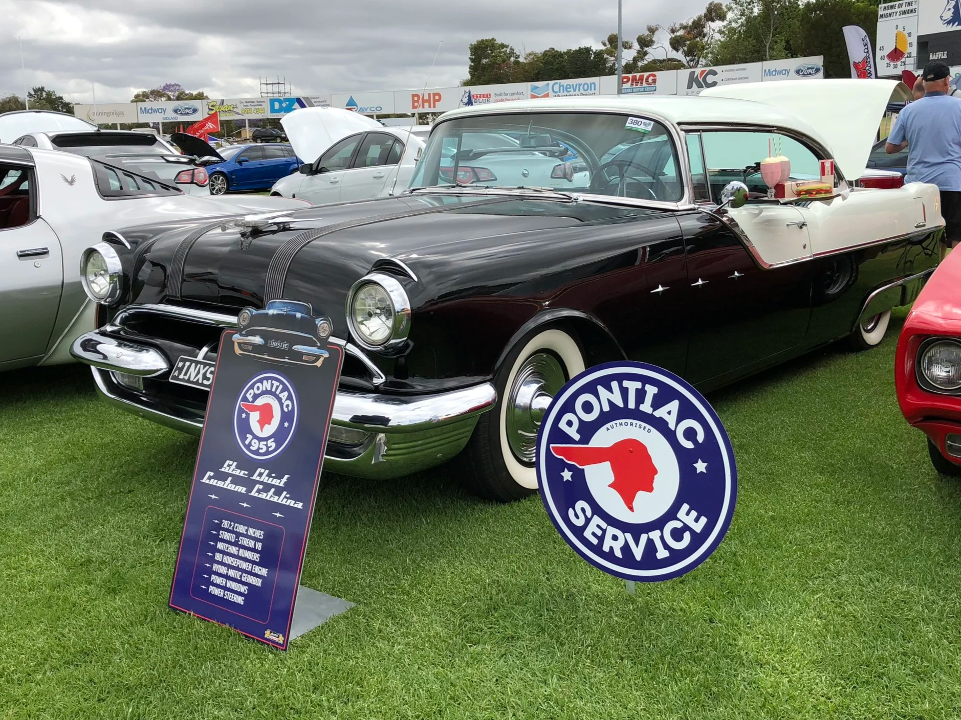 A vintage black and white classic Pontiac car at a car show, with signs and other cars in the background, and a cloudy sky overhead.