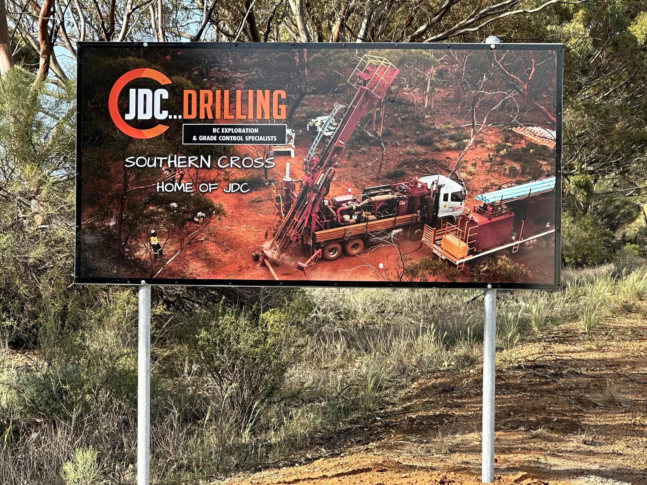 A large outdoor sign with a background image of a drilling operation in a dry, wooded area. The sign reads 'JDC...DRILLING' with smaller text below stating 'RC Exploration & Grade Control Specialists' and 'Southern Cross, Home of JDC'. Road Signage