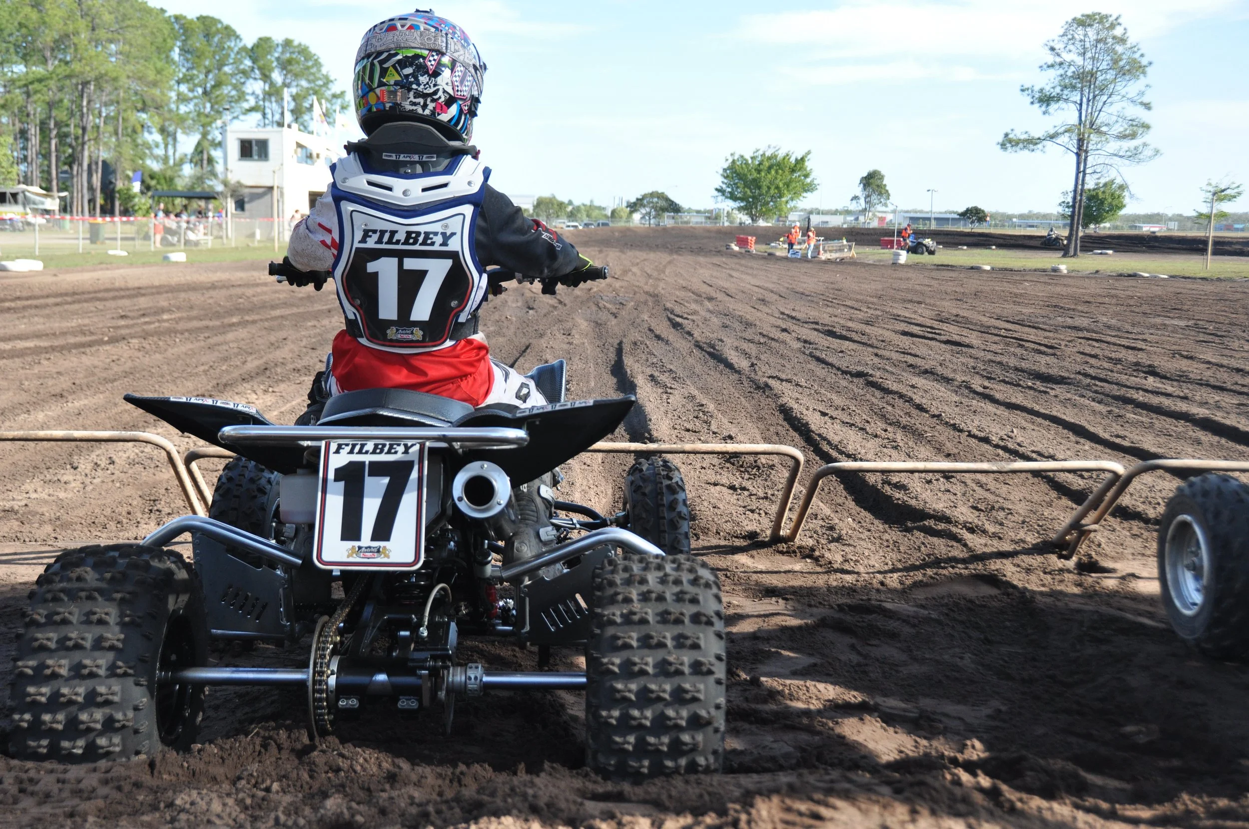 A person riding an ATV on a dirt race track, wearing a helmet and racing gear with the number 17. The track is surrounded by trees and safety barriers, and there are a few officials or workers in the background.
