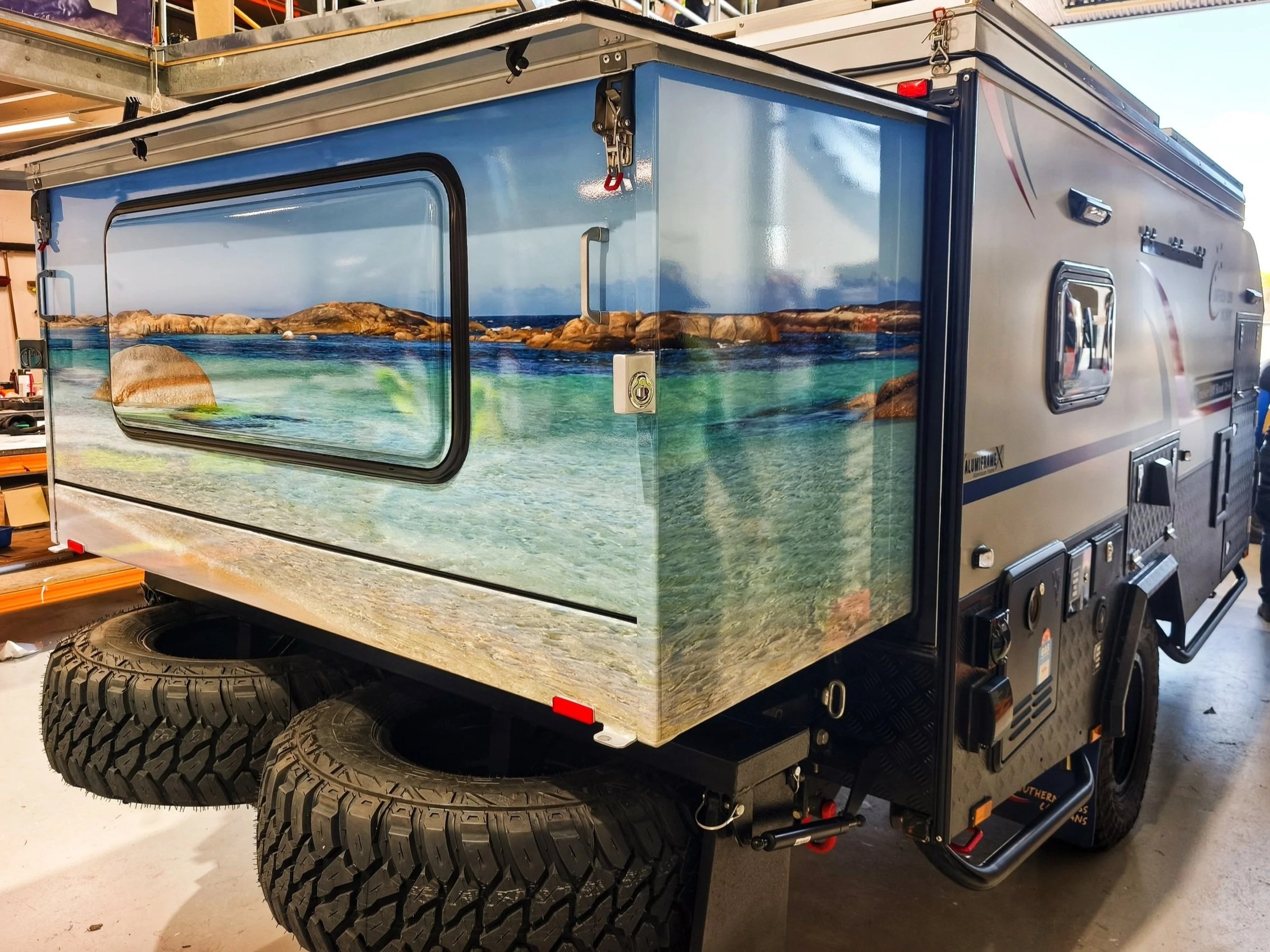 A camper trailer with a scenic landscape mural painted on its side, depicting a beach scene with water, rocks, and mountains.