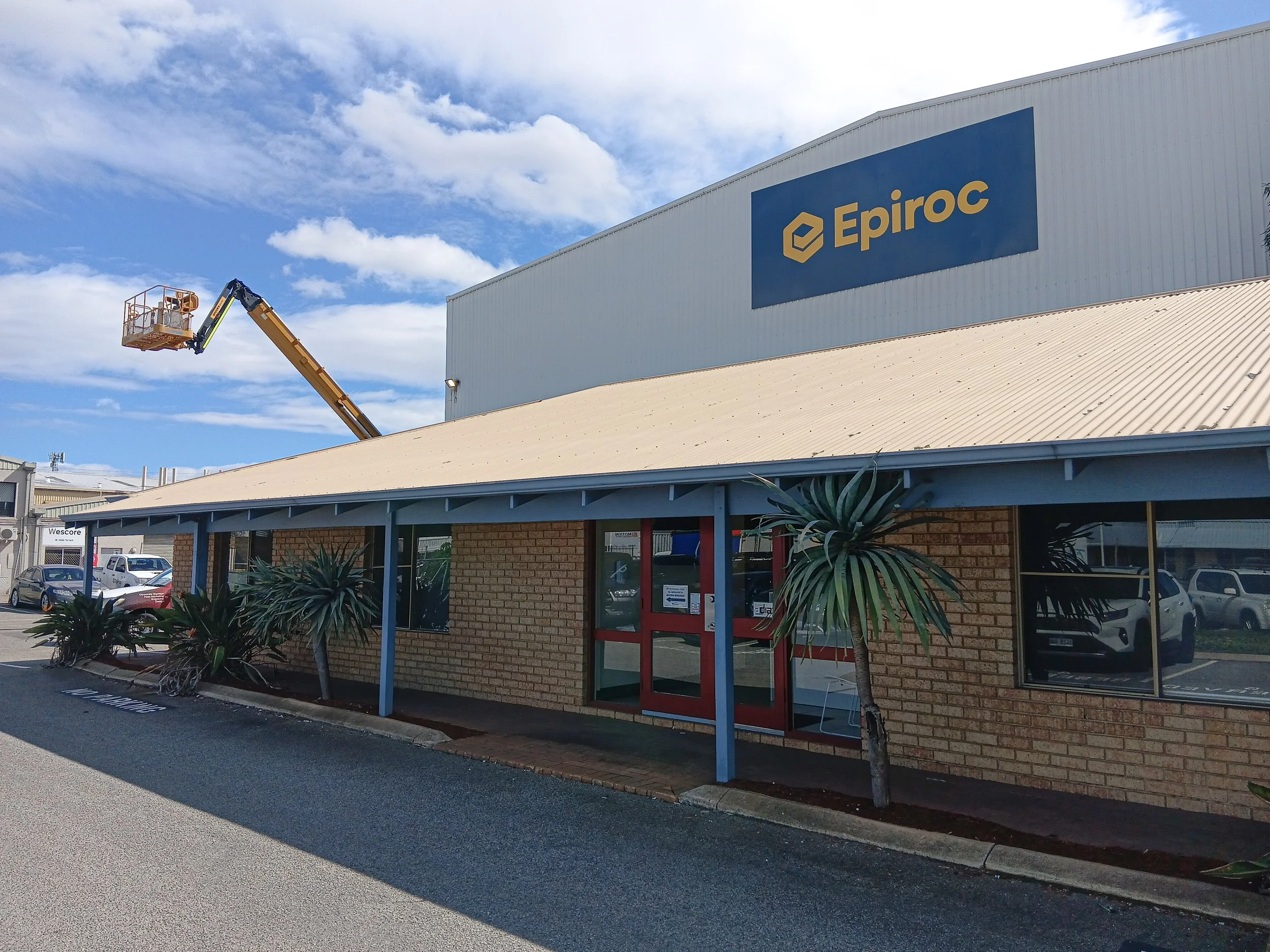 Exterior view of a warehouse or industrial building with a large blue sign displaying the logo and name "Epiroc" fitted high above with a portable lift. Business Signage