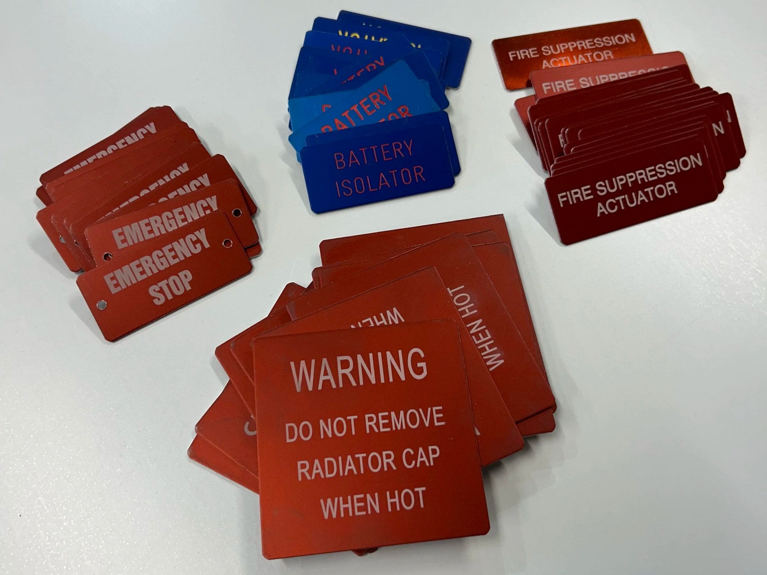 Various red, blue, and orange safety hazard tags including Emergency Stop, Battery Isolator, Fire Suppression Actuator, Warning, Do Not Remove, and When Hot, spread out on a white surface.