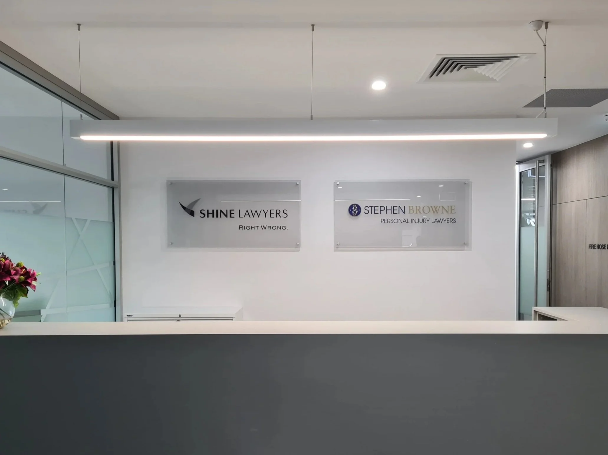 Reception area with glass signage for Shine Lawyers and Stephen Browne Personal Injury Lawyers, white wall, glass partition, ceiling lights, ceiling air vents, flowers on the left side, and a gray countertop visible in the foreground.
