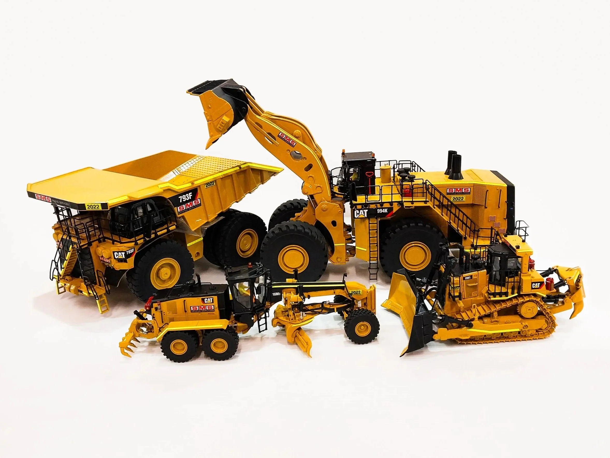 A collection of yellow scale model mining vehicles, including bulldozers, excavators, and dump trucks, arranged on a white background. Custom mining gifts