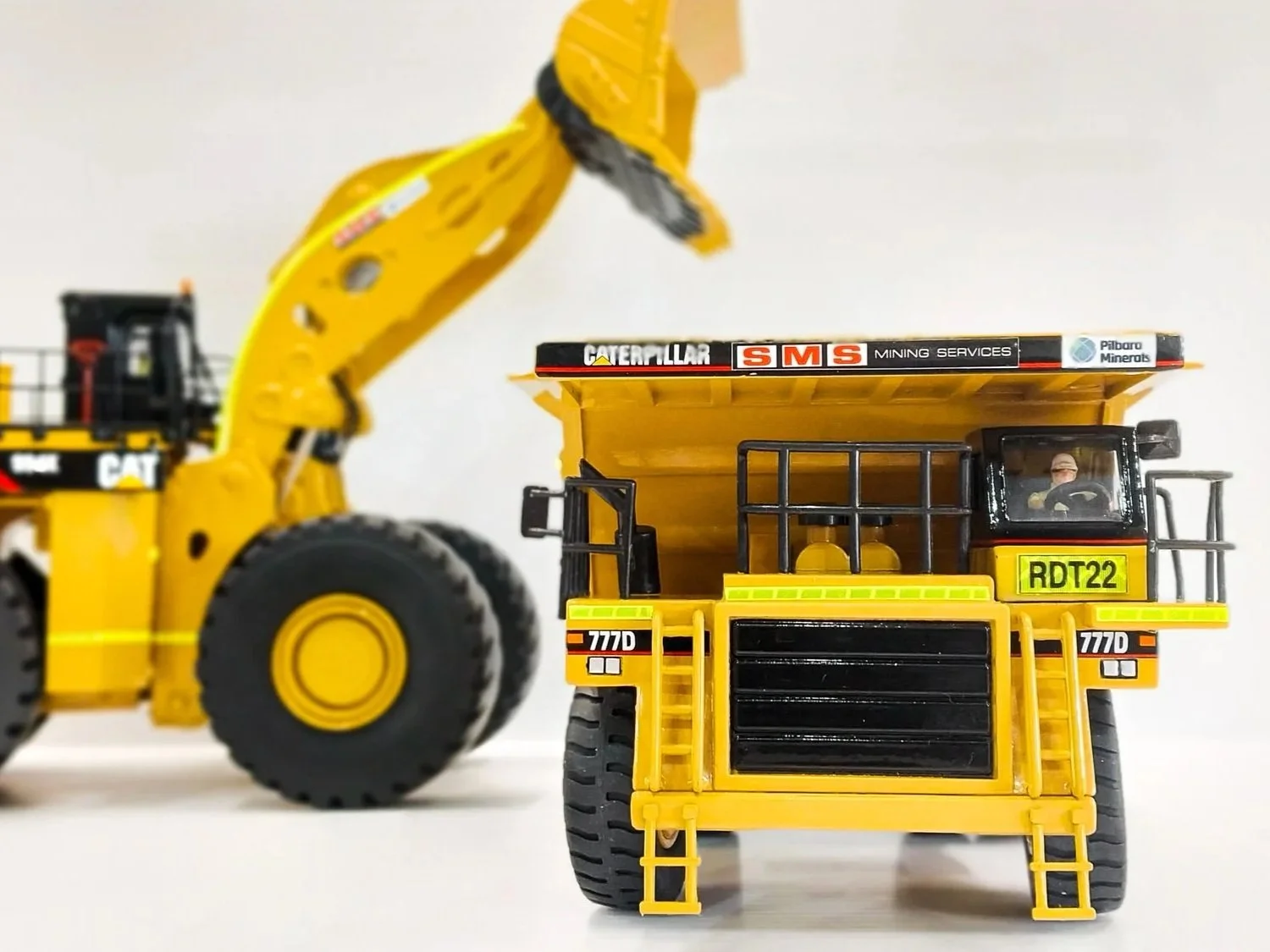 Close-up of scale model mining vehicles, including a dump truck with a driver inside and a front loader with a large bucket, on a plain white background. Custom mining gifts