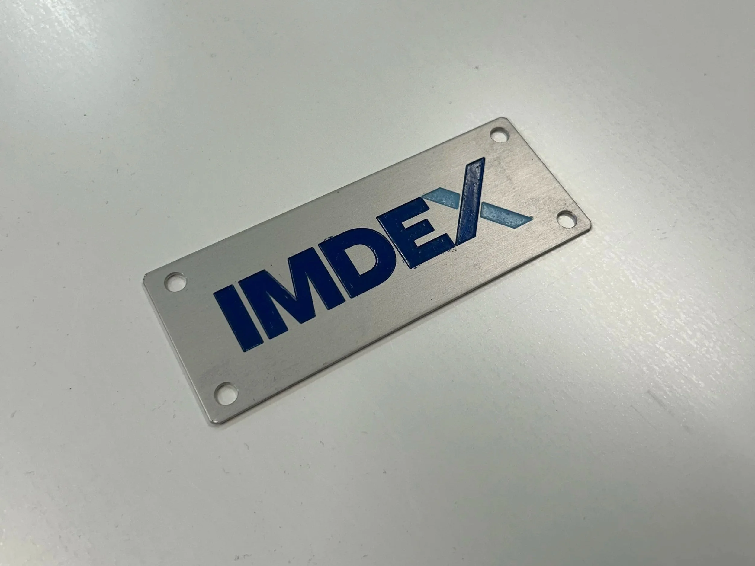 Close-up of a small metal sign with the word 'IMDEX' in blue capital letters, placed on a white surface with four holes in its corners.