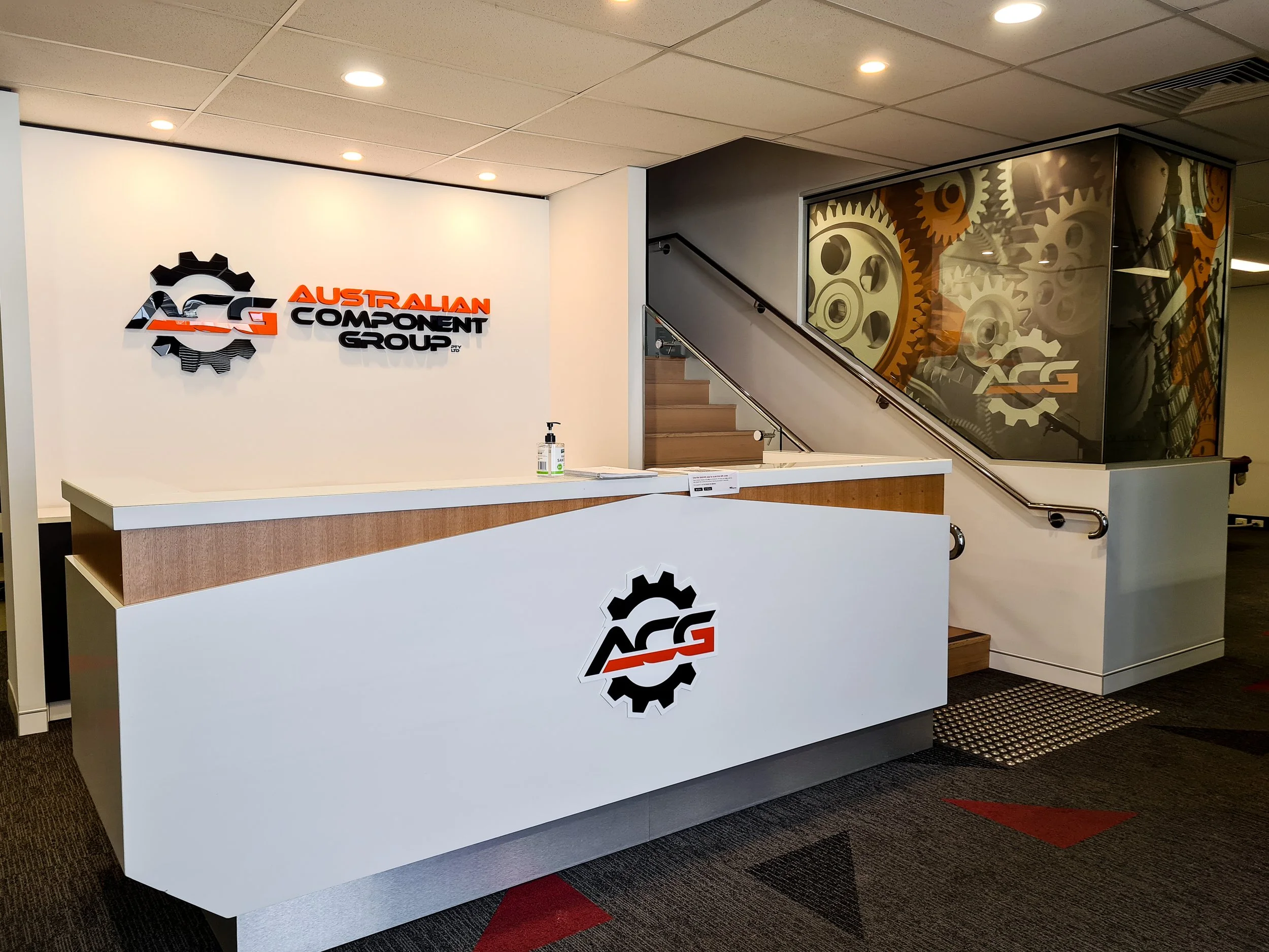 Reception desk at Australian Component Group with a gear logo and a wall featuring gears in the background. Internal front desk business signage