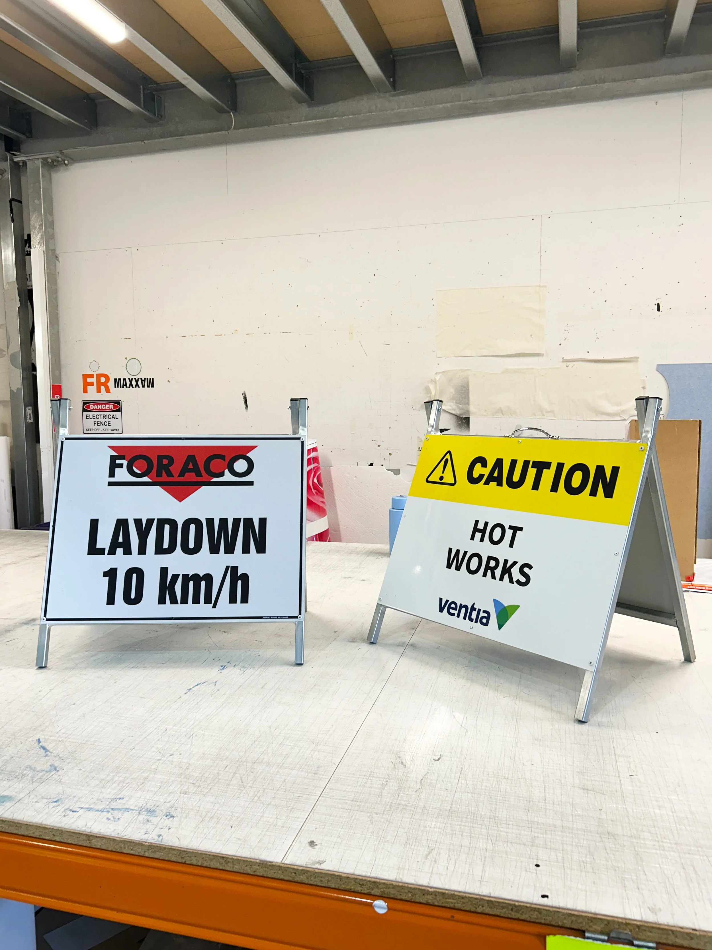 A-Frame signs. One sign reads "LAYDOWN 10 km/h" and another sign reads "CAUTION HOT WORKS." PPE Safety Signage and Regulatory Signage. Portable Signage on High Visibility vinyl